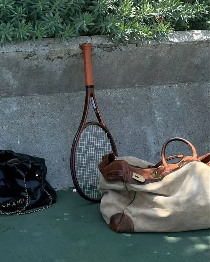 A tennis racket upright on the ground with a bag and a black Chanel handbag nearby, against a concrete wall with greenery above.