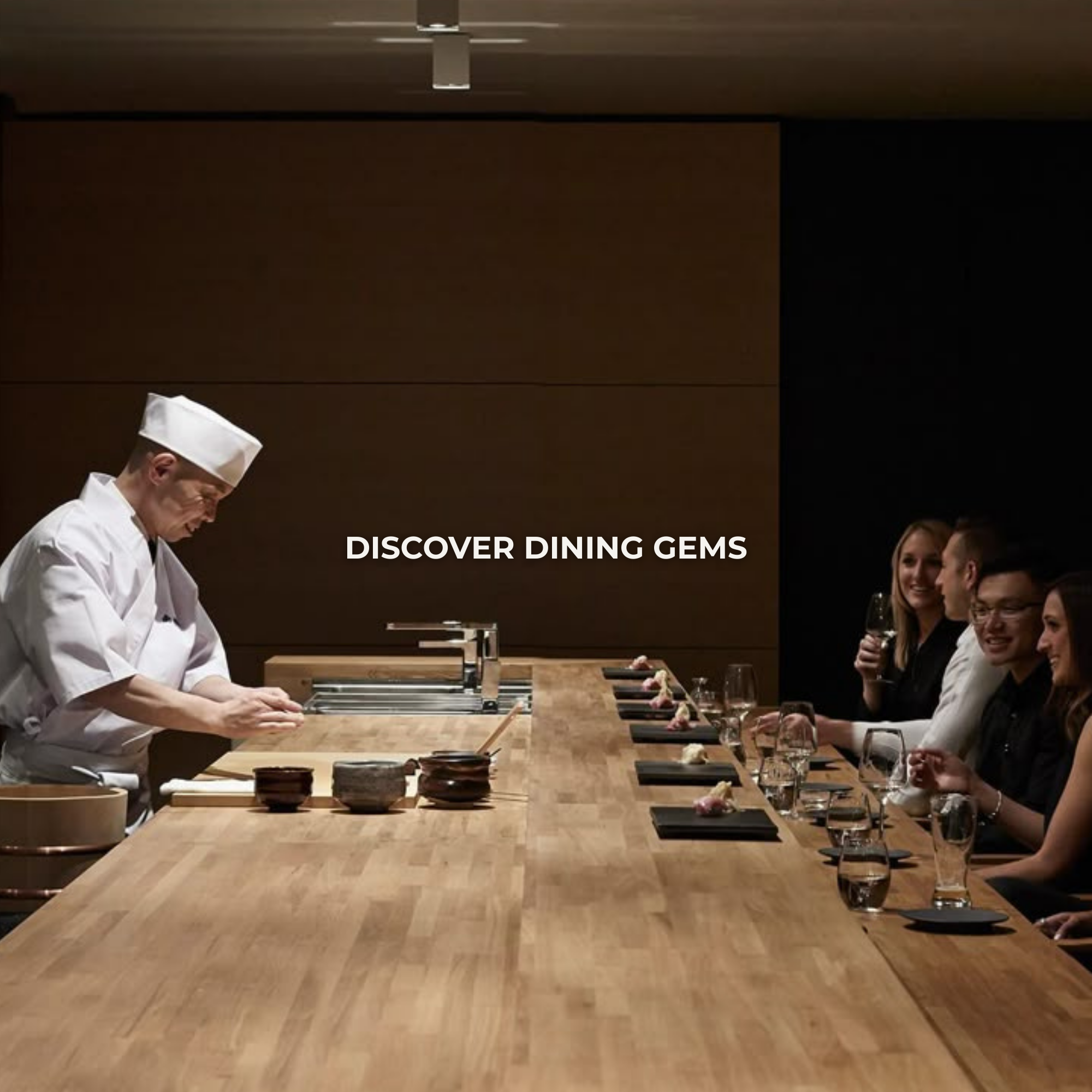 A chef in white uniform and hat preparing food at a wooden counter, with four guests seated on the right side holding glasses and engaging in conversation in a modern restaurant setting.