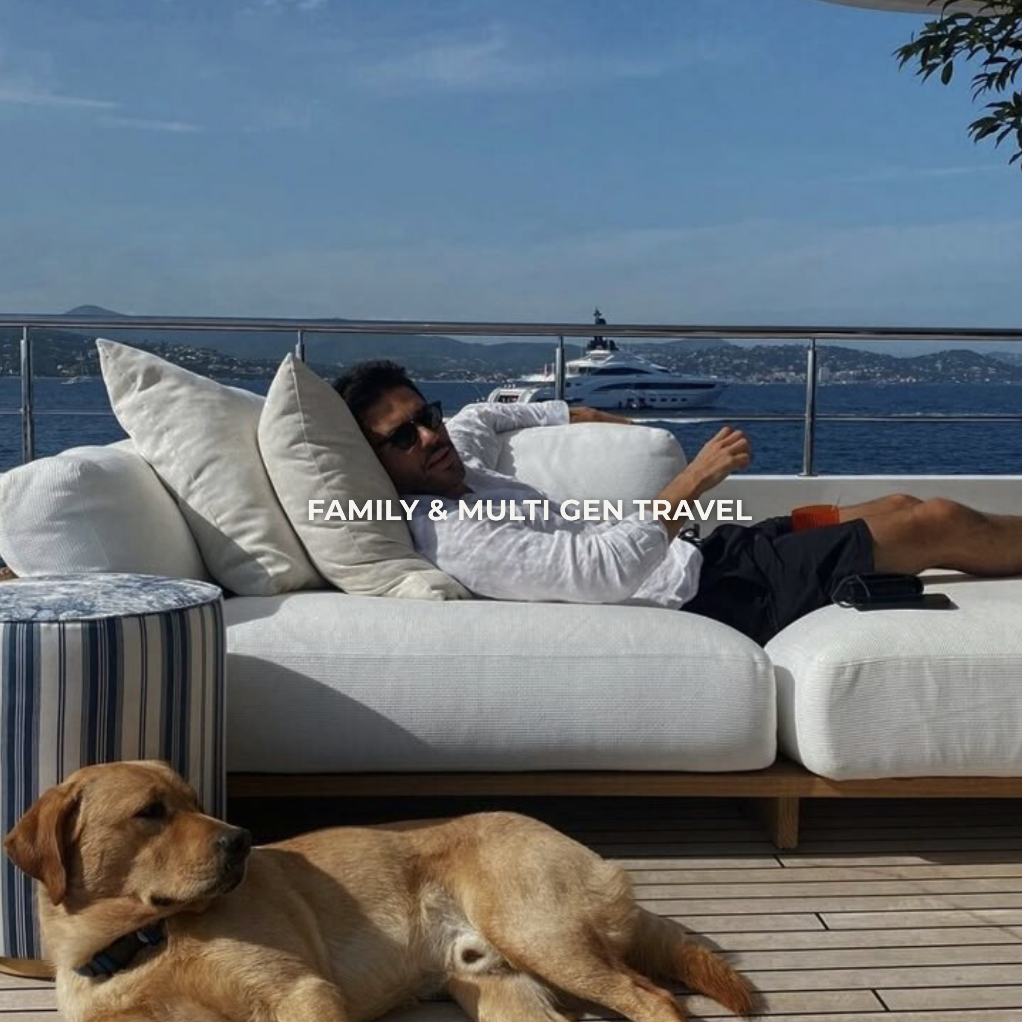 Man relaxing on a sofa with pillows on a yacht, with a dog lying on the deck nearby, and a boat in the background on the water during daytime.