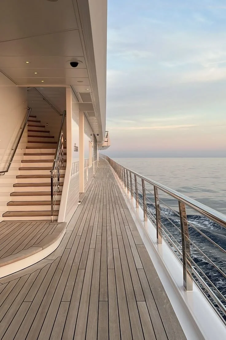 Empty ship deck with wooden flooring, metal railing, and staircase, overlooking calm ocean waters during sunset.