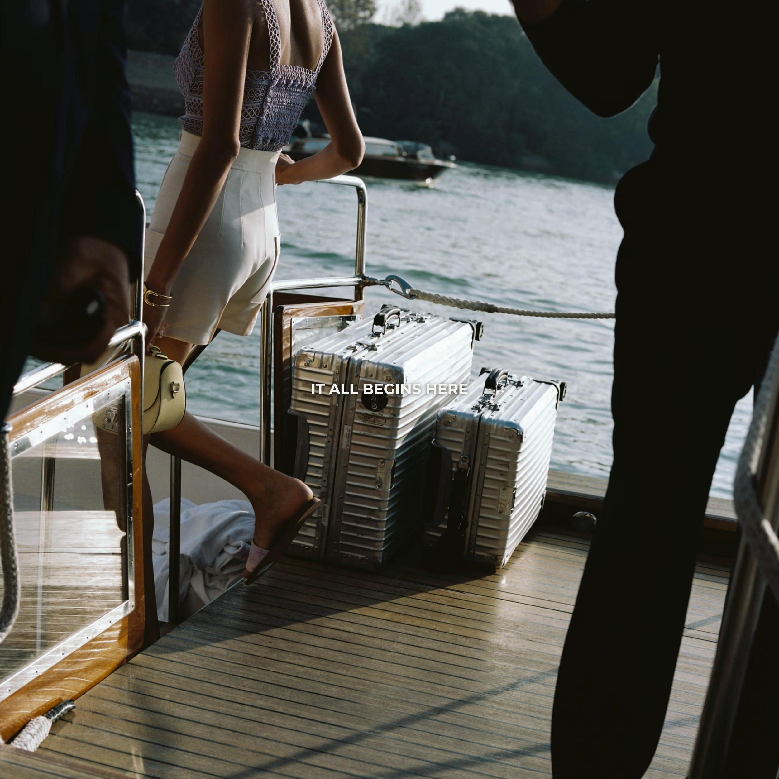 A woman on a boat, wearing a lace top and white shorts, standing near luggage as the boat sails on a body of water with trees in the background. The text on the image reads, 'IT ALL BEGINS HERE.'