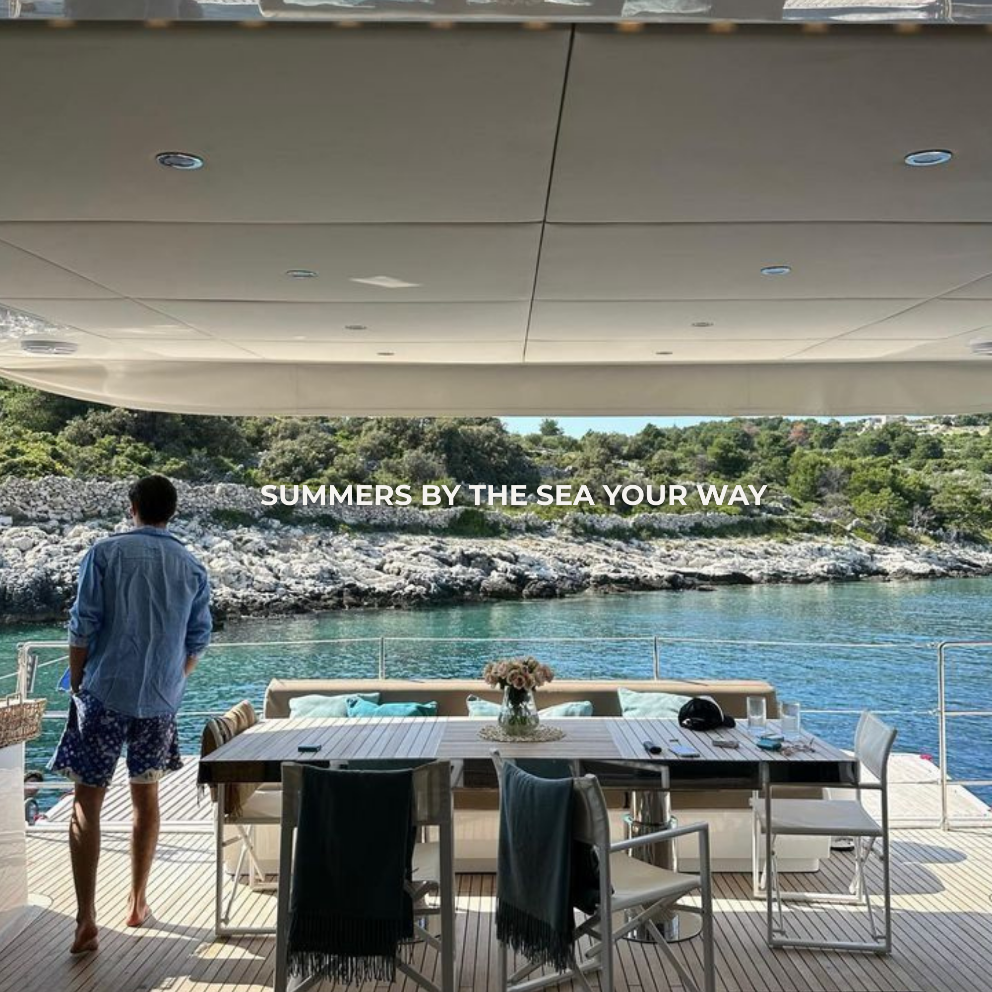 A man stands on the deck of a yacht, overlooking rocky coastline and water, with a dining table and chairs in the foreground.