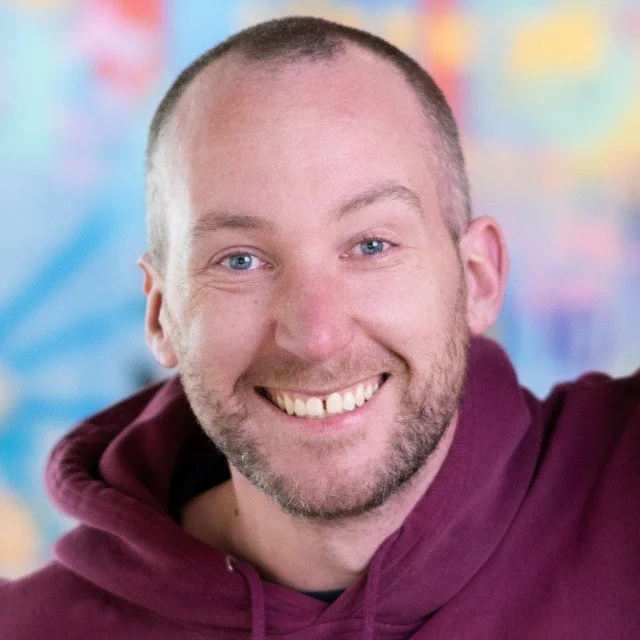 A smiling man with short hair and a beard, wearing a maroon hoodie, in front of a colorful blurred background.
