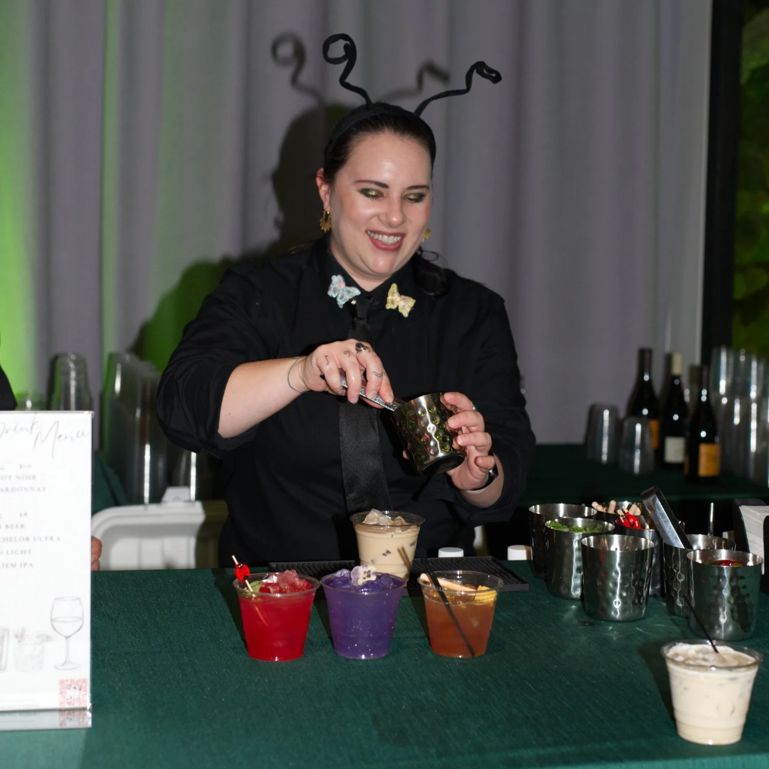 A woman dressed as a bumblebee bartender, wearing antenna headband and butterfly jewelry, preparing colorful cocktails behind a bar with various ingredients.