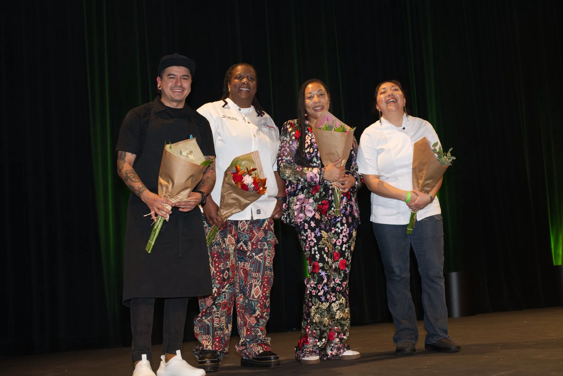 Four people standing on a stage, holding bouquets of flowers, with a dark curtain background, celebrating an event.