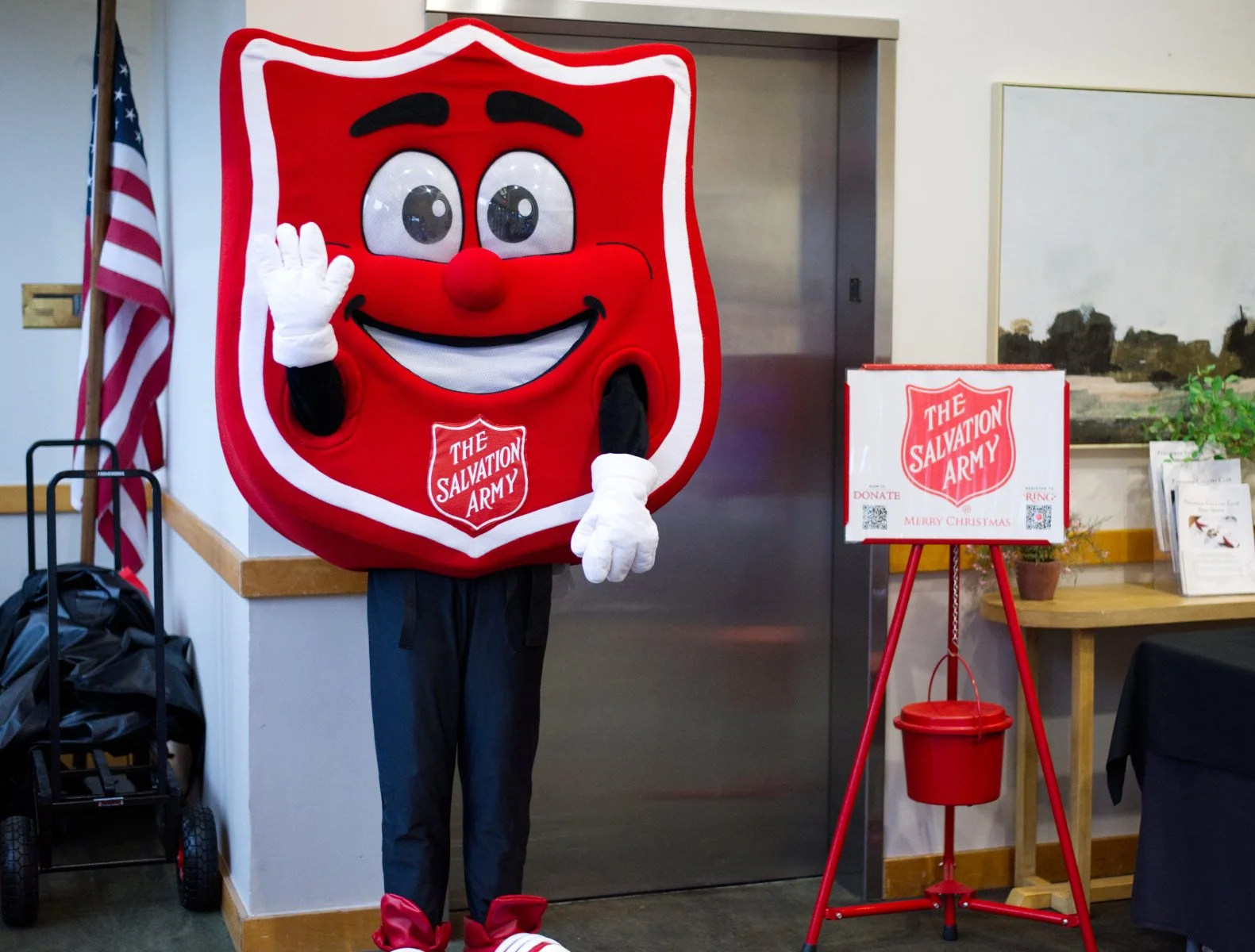 Person in a Salvation Army costume with white gloves, black pants, and red shoes, waving inside a room with American flag and Salvation Army donation stand.