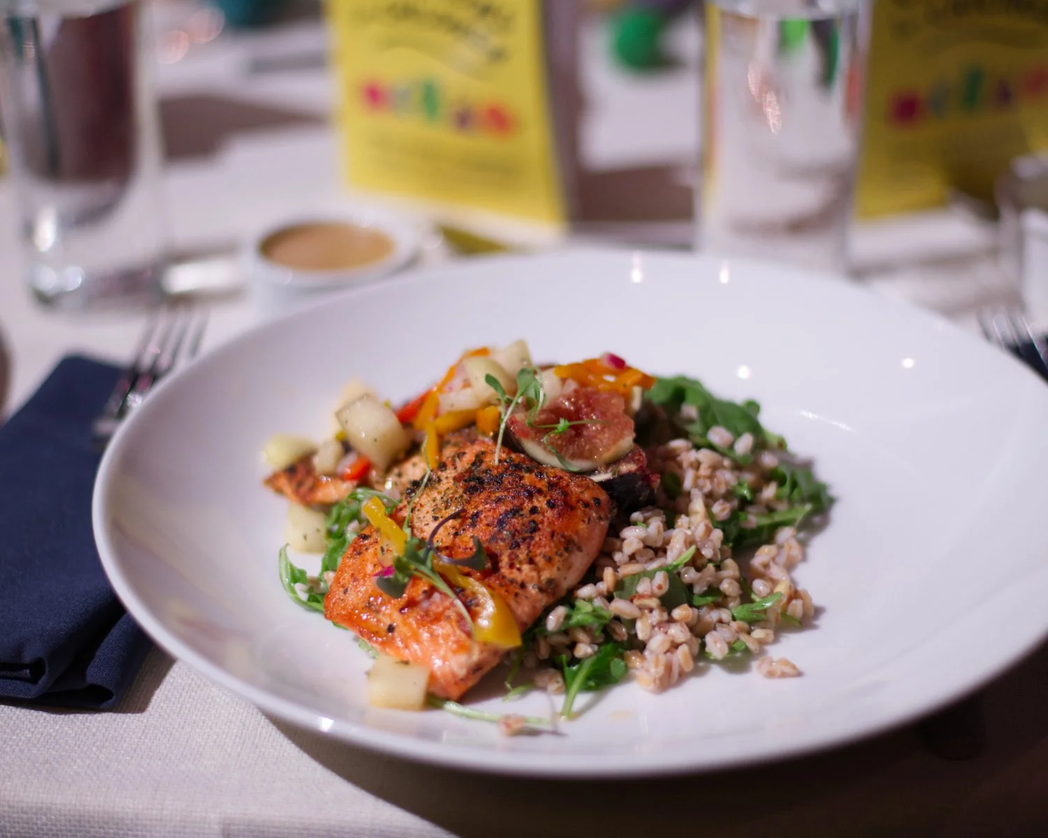 Plate of cooked salmon, grains, and vegetables in a white dish on a table with glasses and drinkboxes in the background.