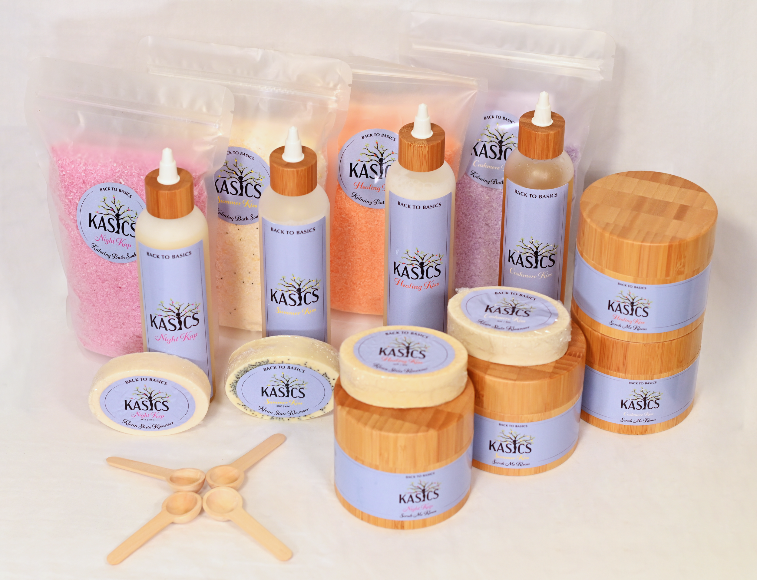 Assorted bath and skincare products including powders, lotions, and scrubs with labels from the brand 'KASICS', displayed with small wooden spoons for sampling, set against a plain background.