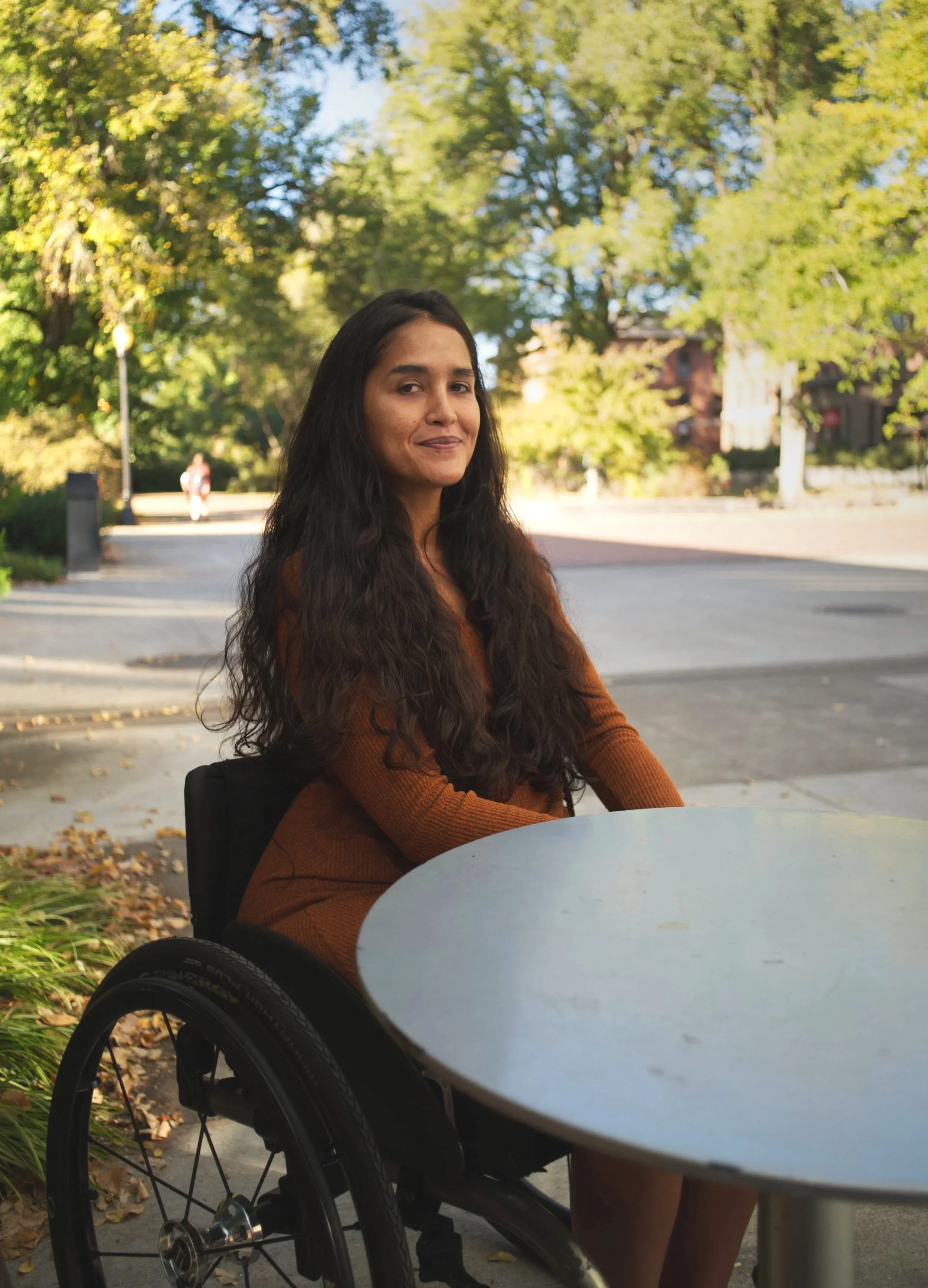A woman with long dark hair sitting at an outdoor table in a wheelchair, wearing a brown sweater, with trees and a sidewalk in the background during daytime.