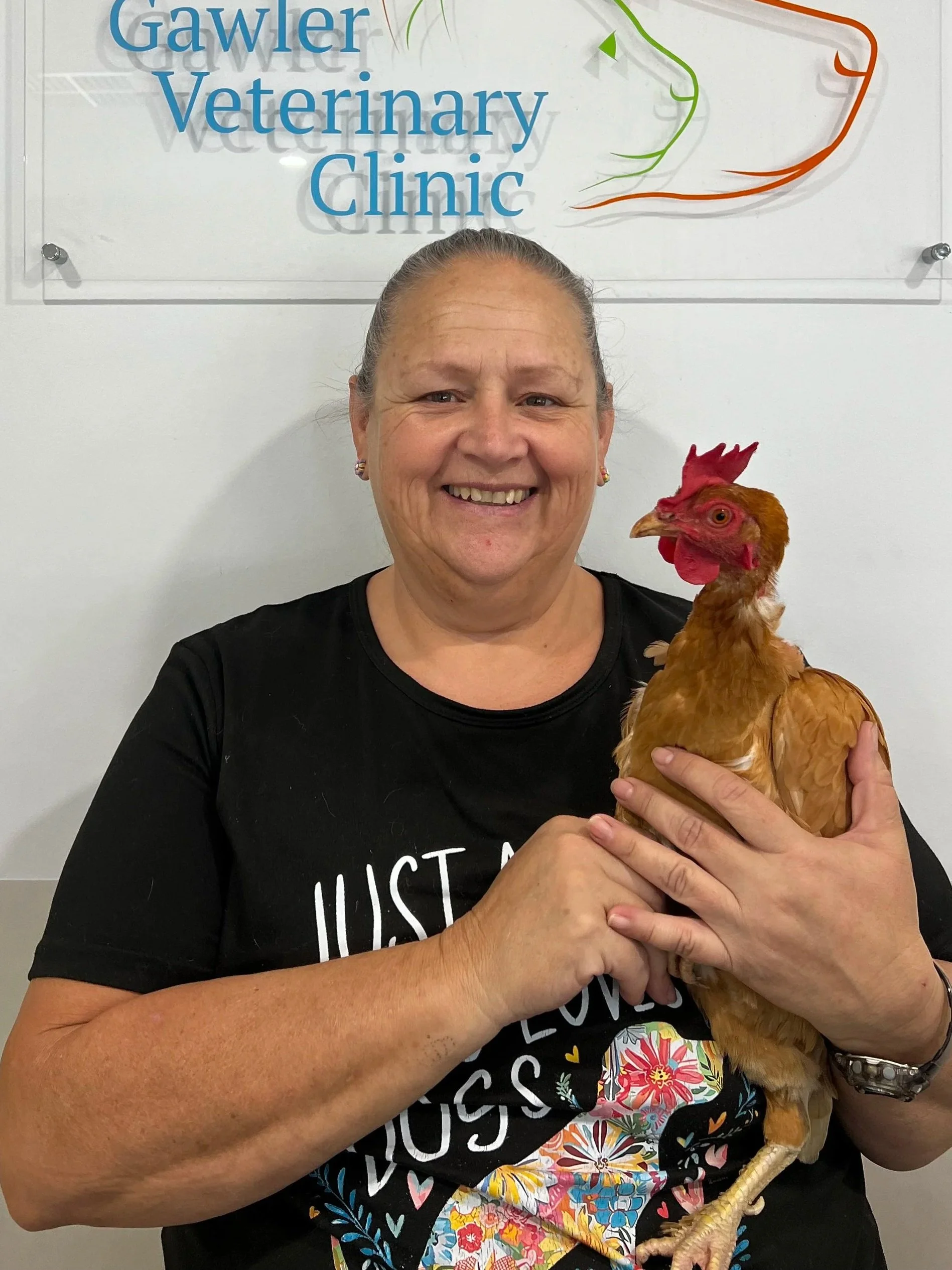 A woman smiling and holding a chicken inside a veterinary clinic with a sign that reads "Gawler Veterinary Clinic" behind her.