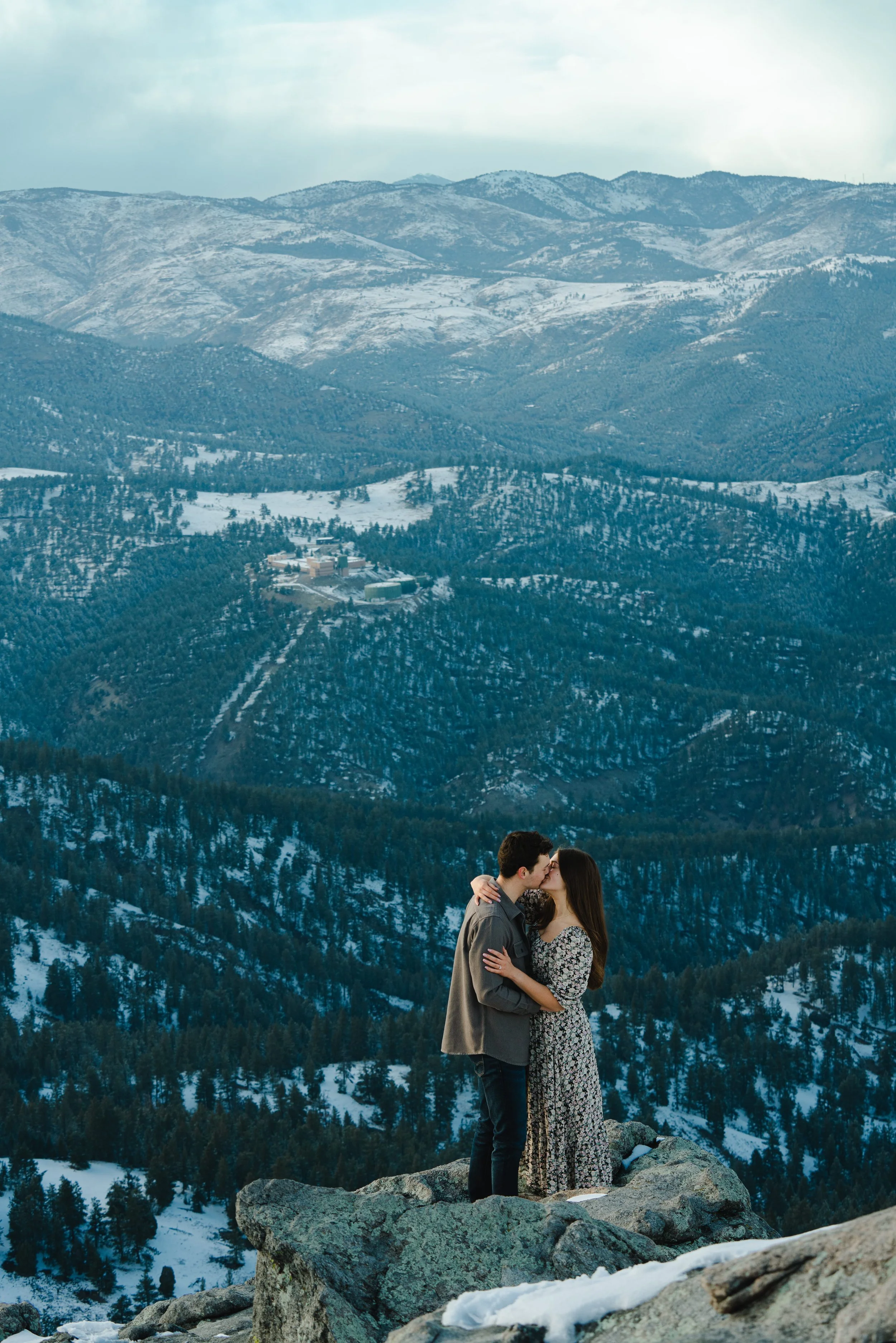 A newly engaged couple embracing over a snowy vast mountain overlook.  The mountains stretch to the top of the frame and seem miles away.