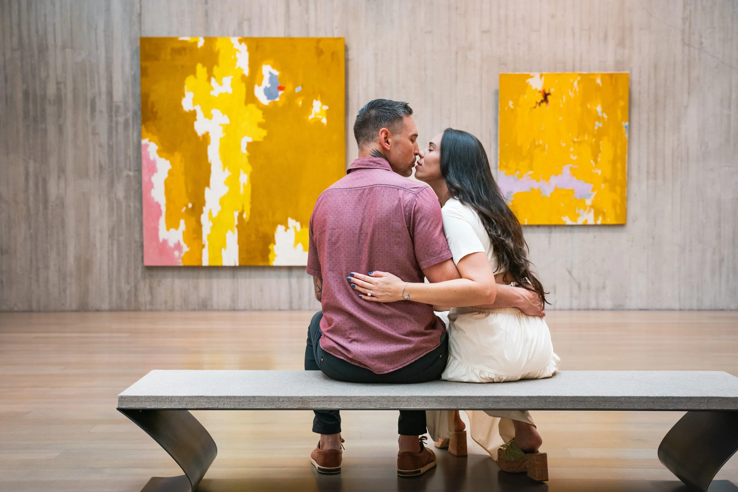 A couple sit in the Clyfford Stills Museum in Denver Co.  They are framed in between two yellow canvases in front of them.  They are inches away from kissing with the female's left hand on his back, showing her engagement ring.