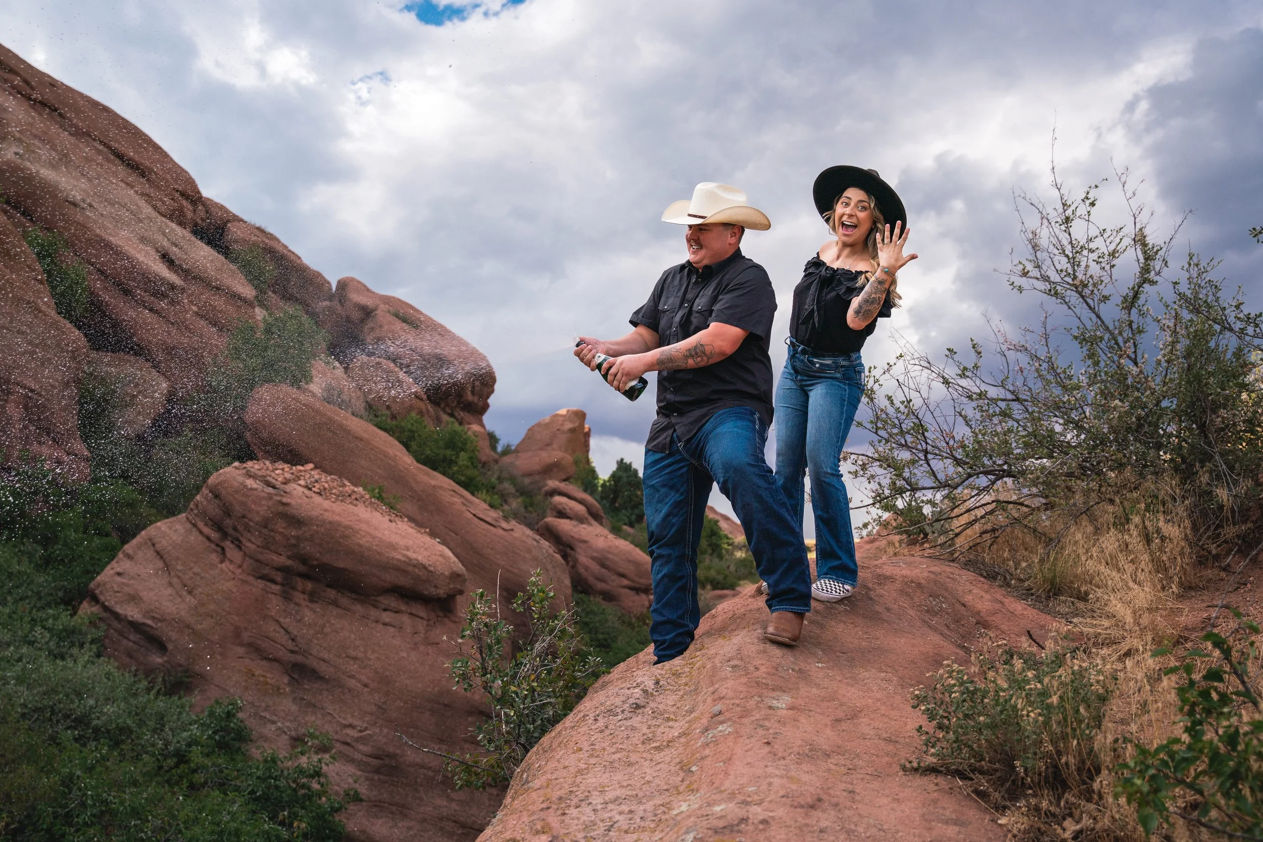 A couple laughing and smiling on their engagement photo session.  They are in western attire, both wearing cowboy hats while the woman is flashing her ring at the camera and the man is blasting a bottle of champagne into a slot canyon in Red Rocks.