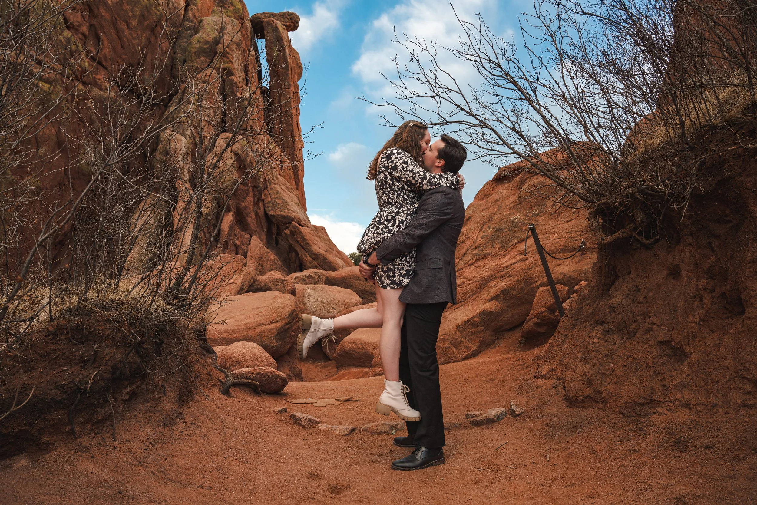 A couple embrace while the man holds his newly engaged fiancé in the air.  They are hugging and kissing with a bright blue sky in the background and the stark contrasting red rocks from Garden of the Gods, Colorado.