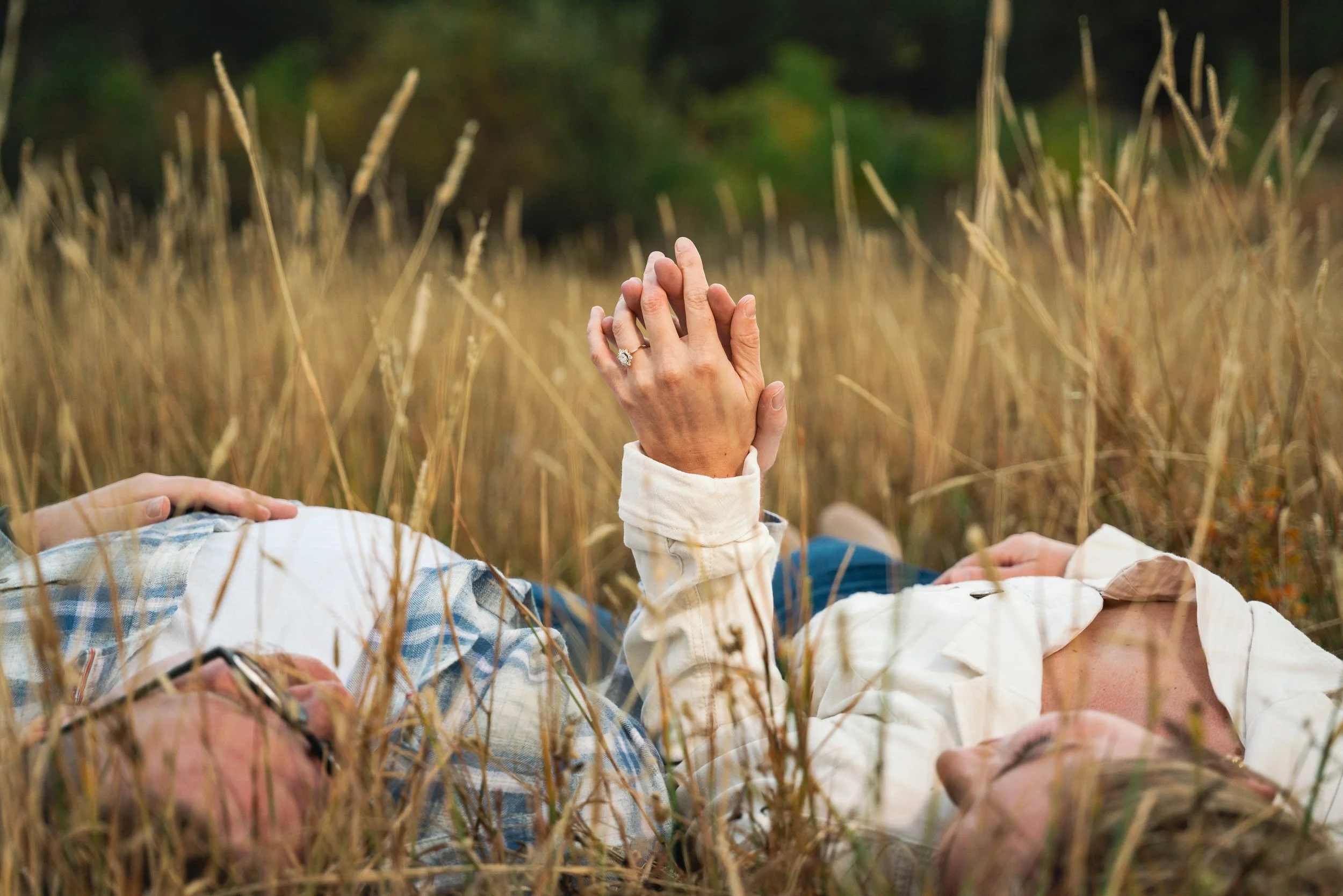 A couple lay in the grass, looking at each other.  The main focus in the photo is their hands slightly held up from the ground, and beginning to embrace.  The woman's engagement ring is in the dead center of the photo and in full focus.