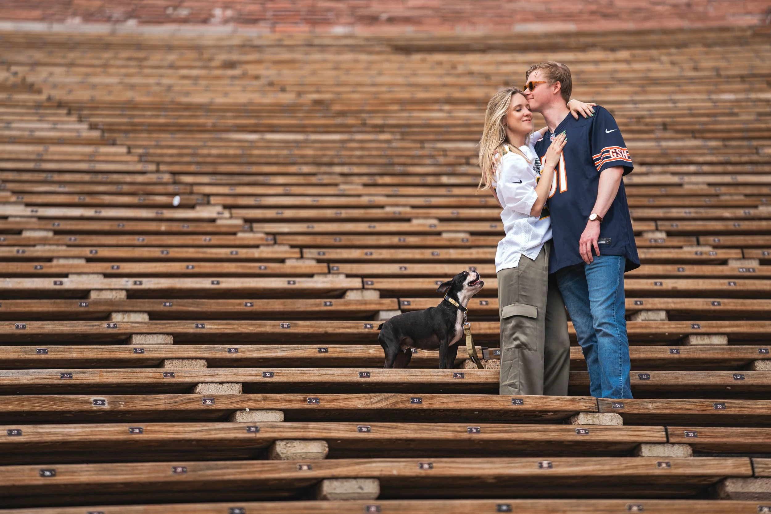 A couple gently embracing on the steps of Red Rocks Amphitheater in Morrison, Colorado.  The pattern of the chairs create an alluring diagonal shape in the image.  Their dog, a boston terrier happily looks up at them while they hug.