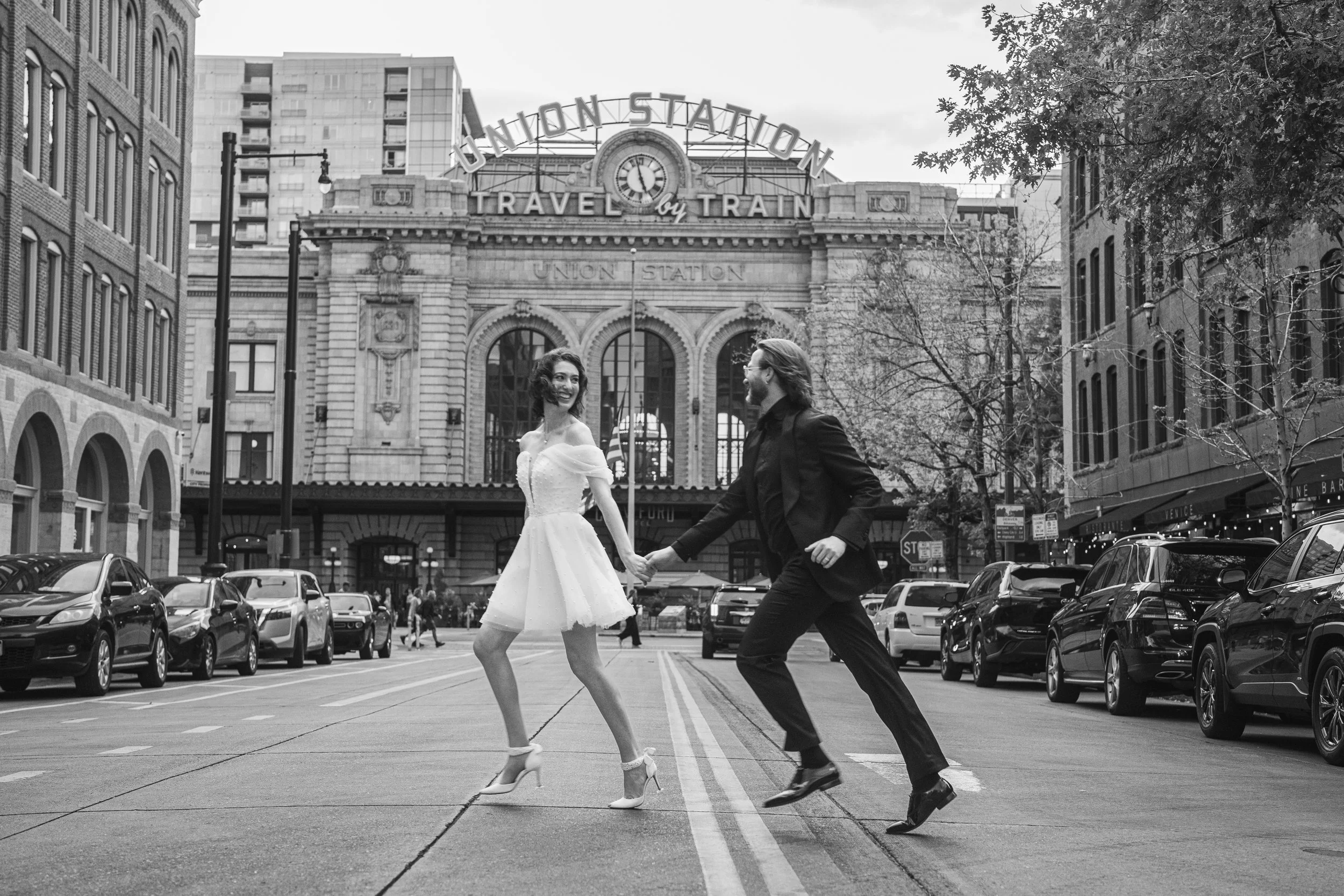 A couple crossing in front of Union Station Train Station in Downtown Denver after a courthouse elopement.  The groom looks back at his wife smiling, hand in hand, after getting married.