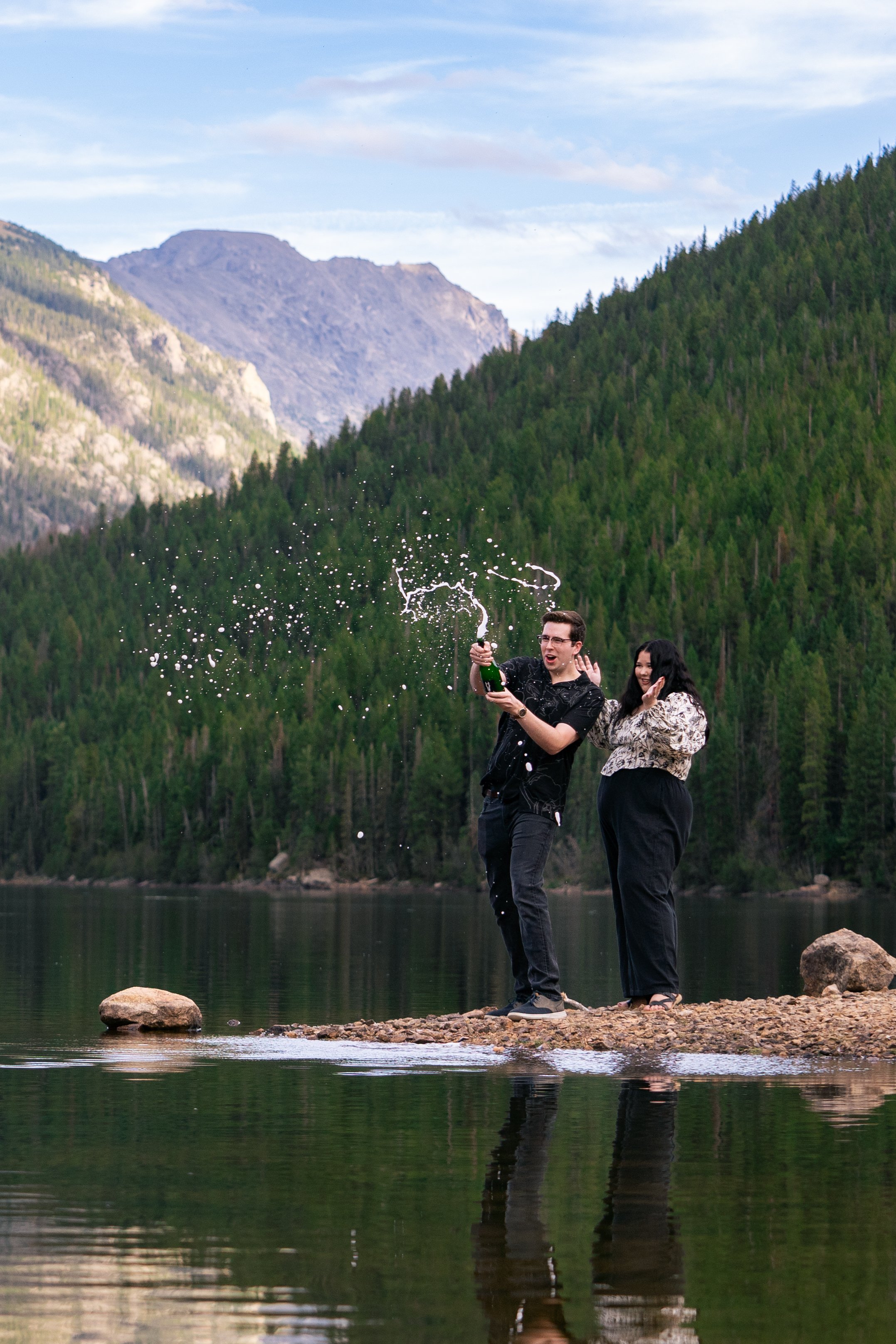 A newly engaged couple pop a bottle of champagne towards a lake at a remote destination.  This shoot was deep in the Colorado mountains, and the couple is completely alone, other than the photographer of course!