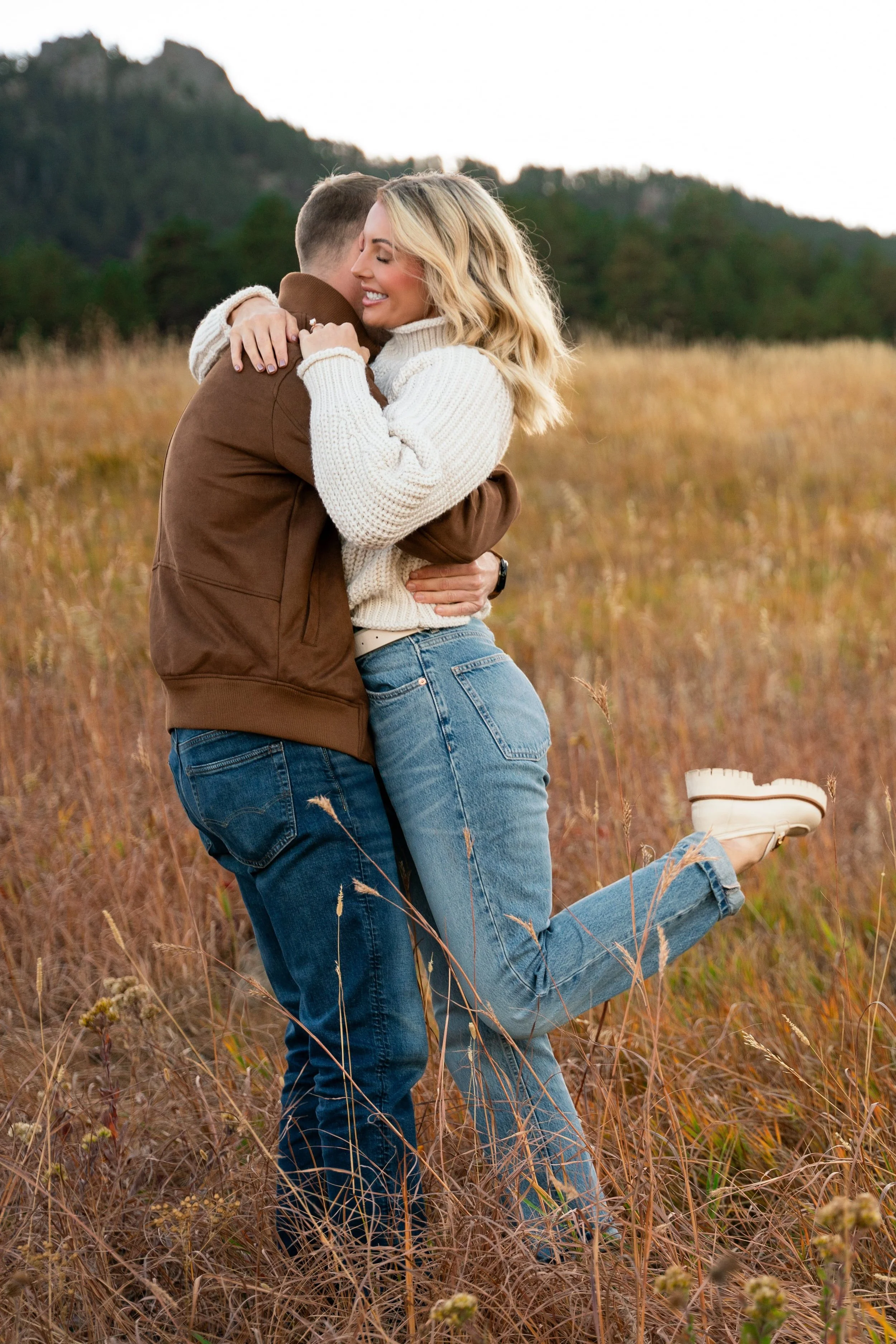 A couple hug after engagement, with the blonde fiancé kicking her back leg up.  They are in a field of yellow tall grass in Boulder, Colorado.  The bride has a big smile and her eyes are closed as she leans into her partner.