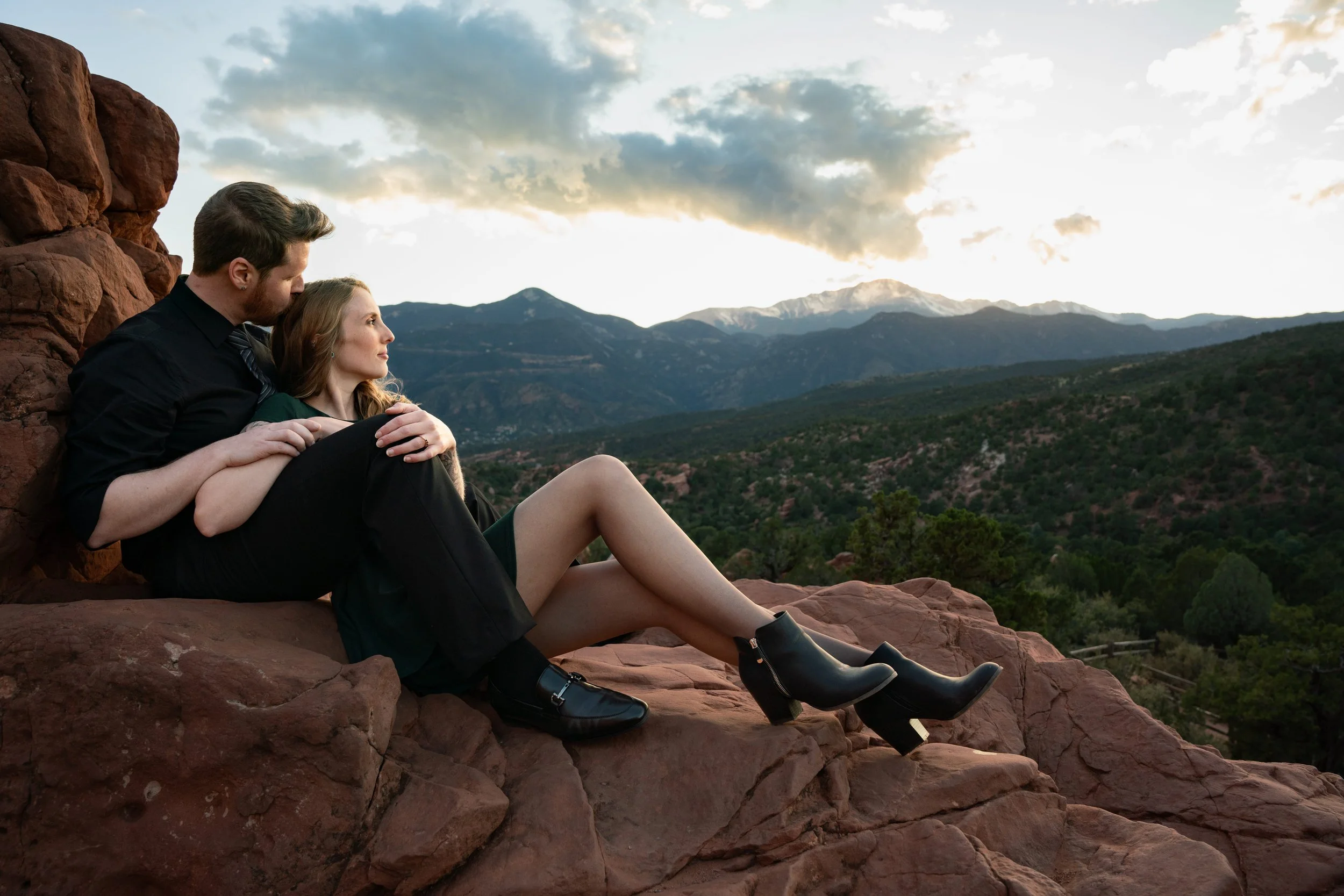 A editorial style photo during a sunset at Garden of the Gods in Colorado Springs.  The couple looks off at the sunset behind the mountains, the man kissing his fiance's head.  Her gaze is elegant, with golden sunlight highlighting her profile.