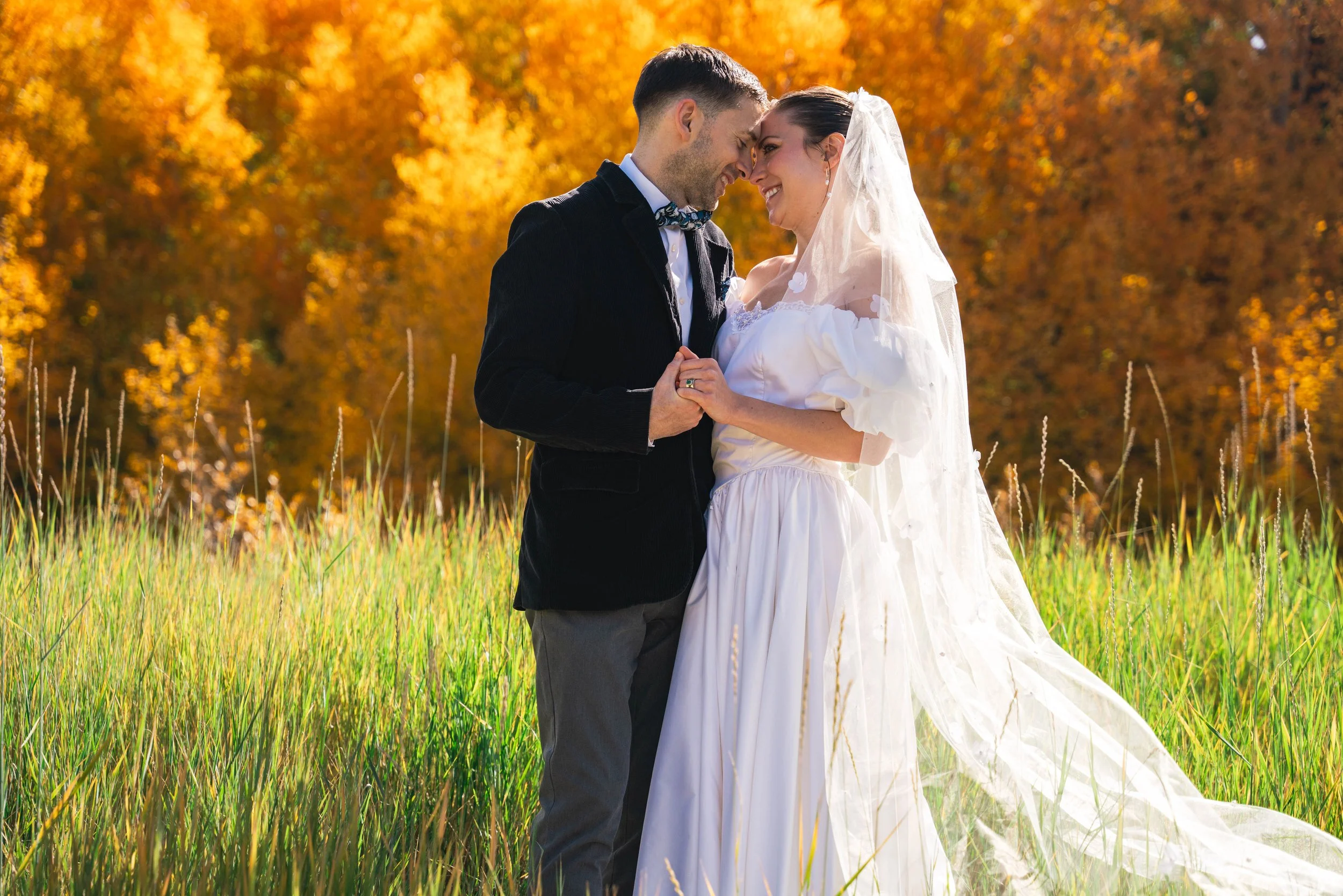 A couple looking into each others eyes and holding eyes, smiling.  They are standing in a green field in front of a vibrantly orange aspen grove in the middle of fall season.  The background contrast is magical for a beautiful elopement.
