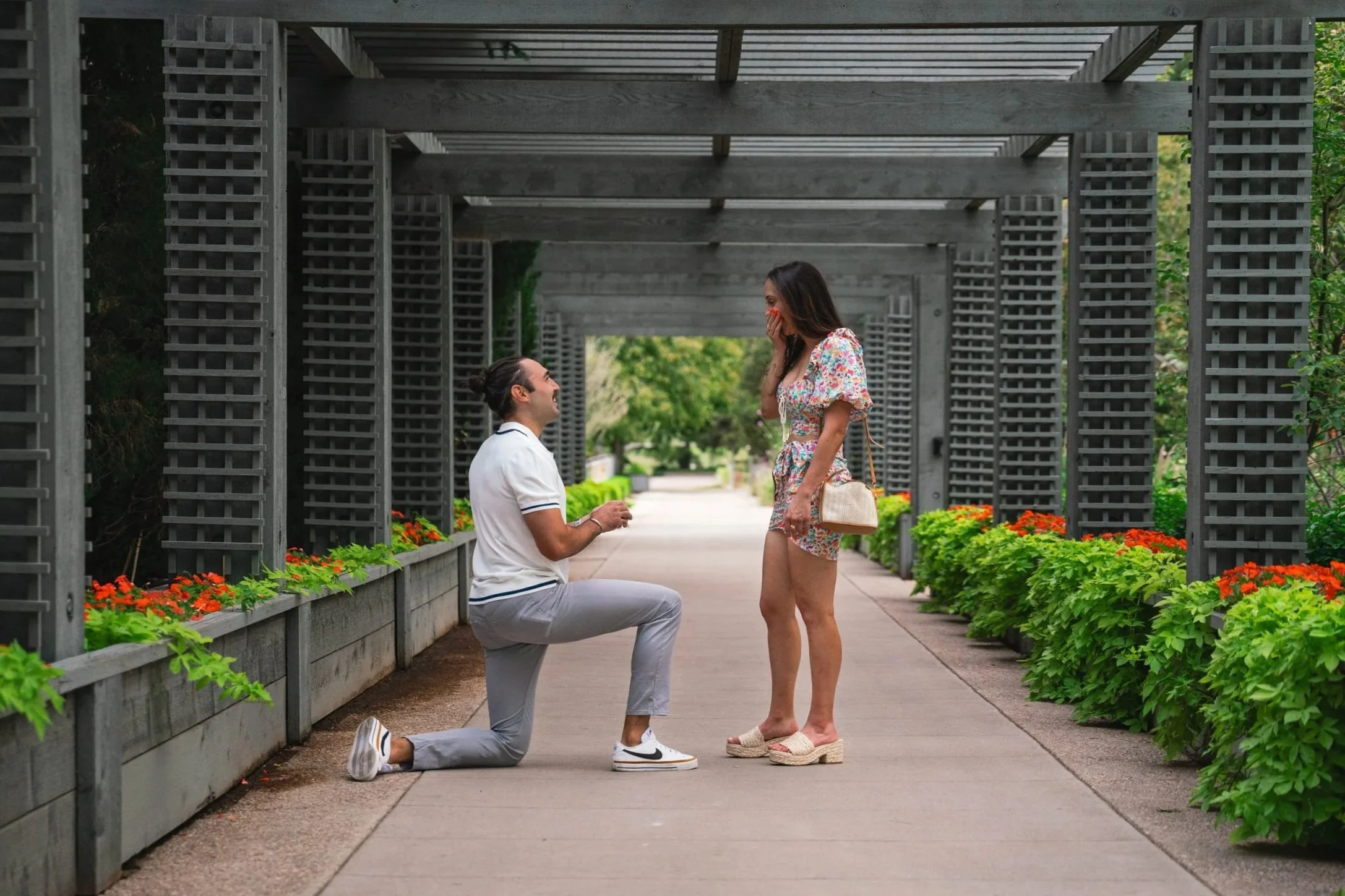 A man is on bended knee, in front of his shocked soon to be fiancé,  Her mouth is agape with her hand covering it.  The moment is candid and authentic.  They are in the botanical gardens in Denver Colorado.