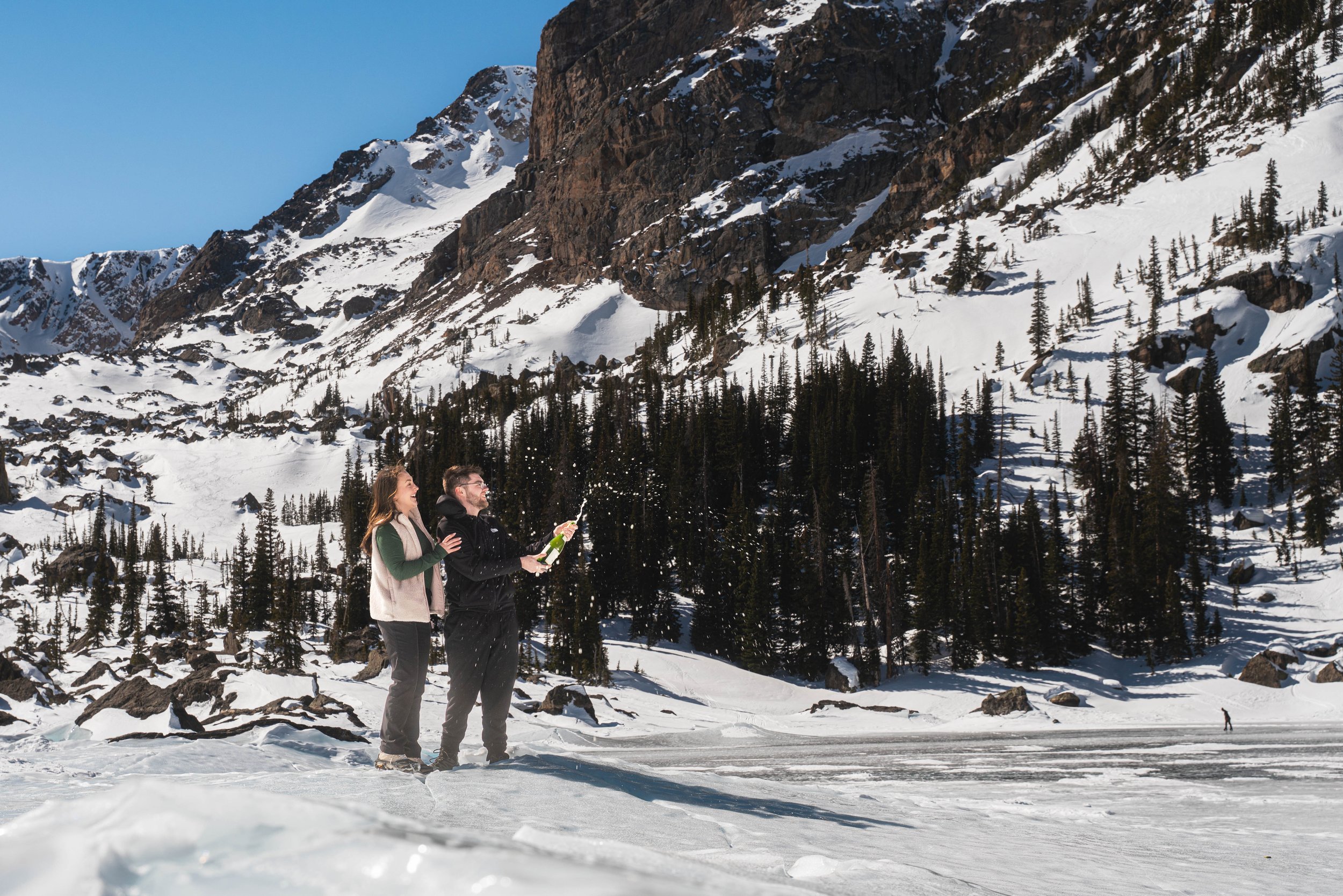 A couple is at the frozen glacier lake, Lake Haiyaha in Rocky Mountain National Park.  After a successful proposal, they are celebrating with champagne on the ice.