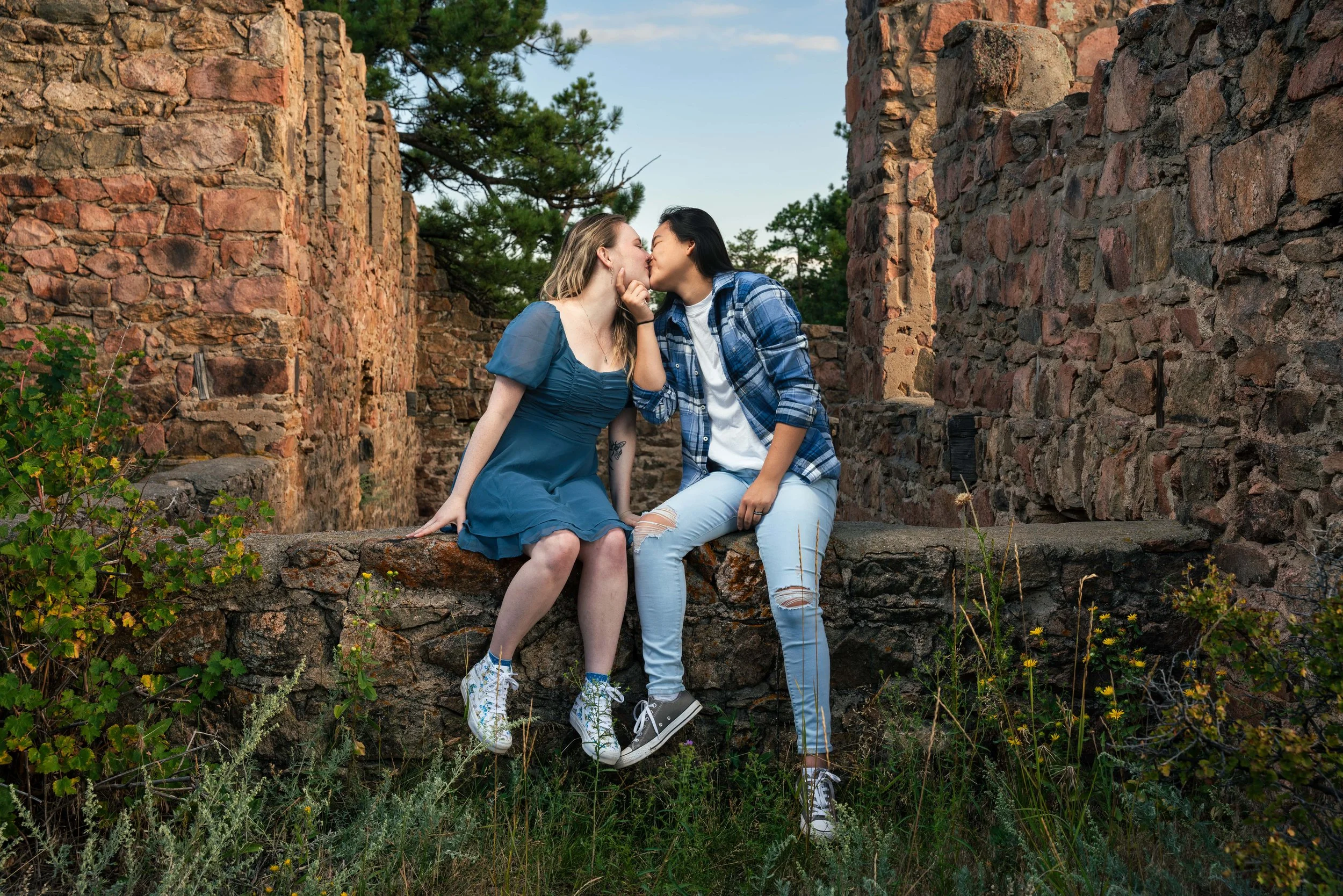 Two women lean in for a kiss at a remote location.  They are sitting on the remains of an abandoned castle.  There is no roof but they are on a stone fence, one of the women holding the other's chin to pull her in for a kiss.  Exceptionally lit photo