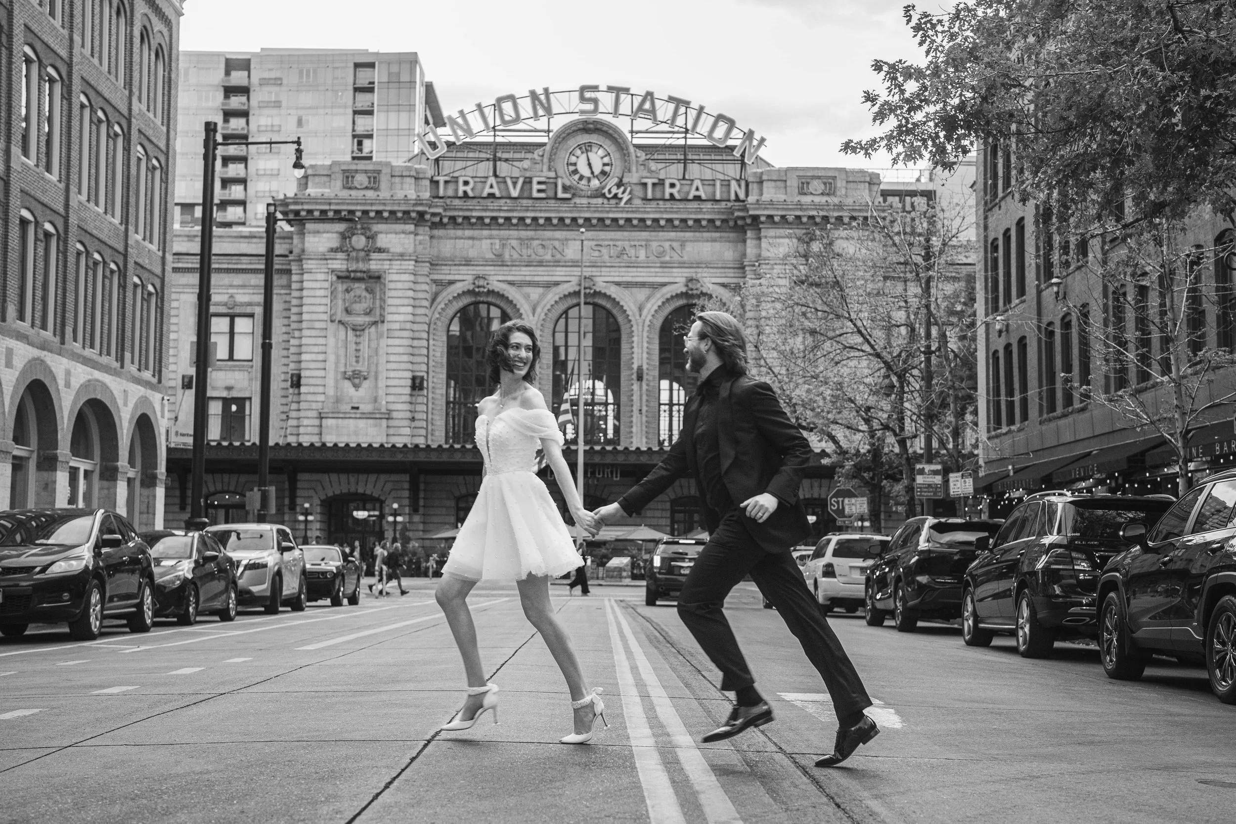 A black and white photo of a couple running across the city street after eloping.  The bride is leading the way in heels and a short wedding dress, holding hands and looking back at her groom.  He is in full stride with her, looking almost like he's 