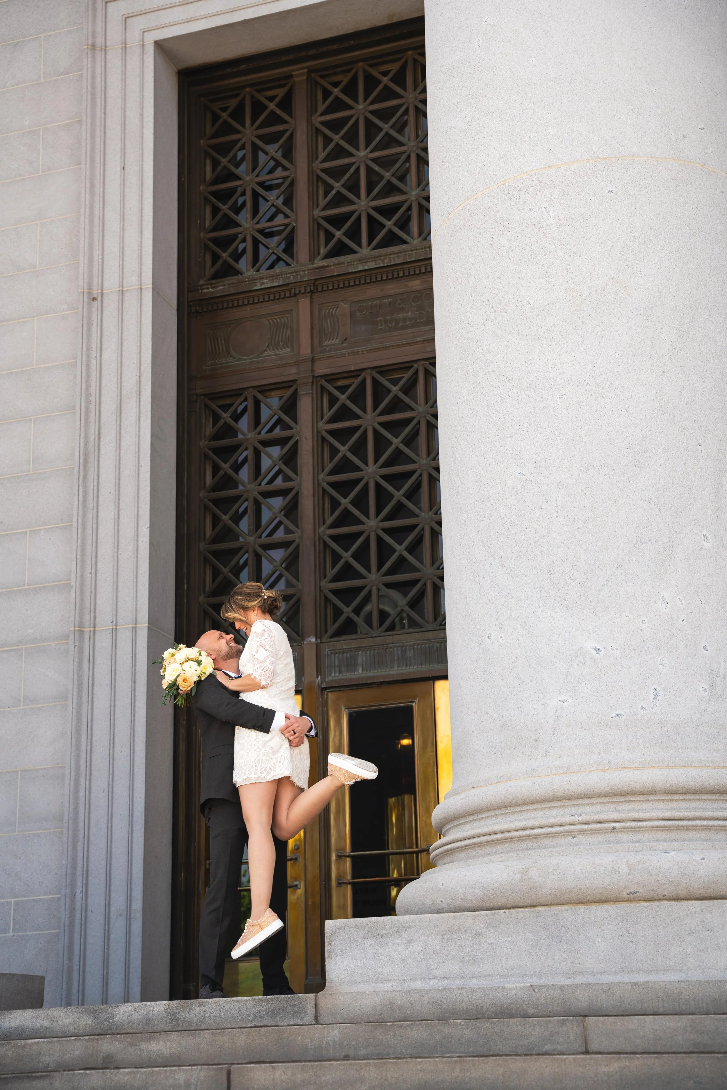 A groom holding his bride up in the air, looking into her eyes.  They are on the steps of a courthouse after their elopement in Denver, Colorado.