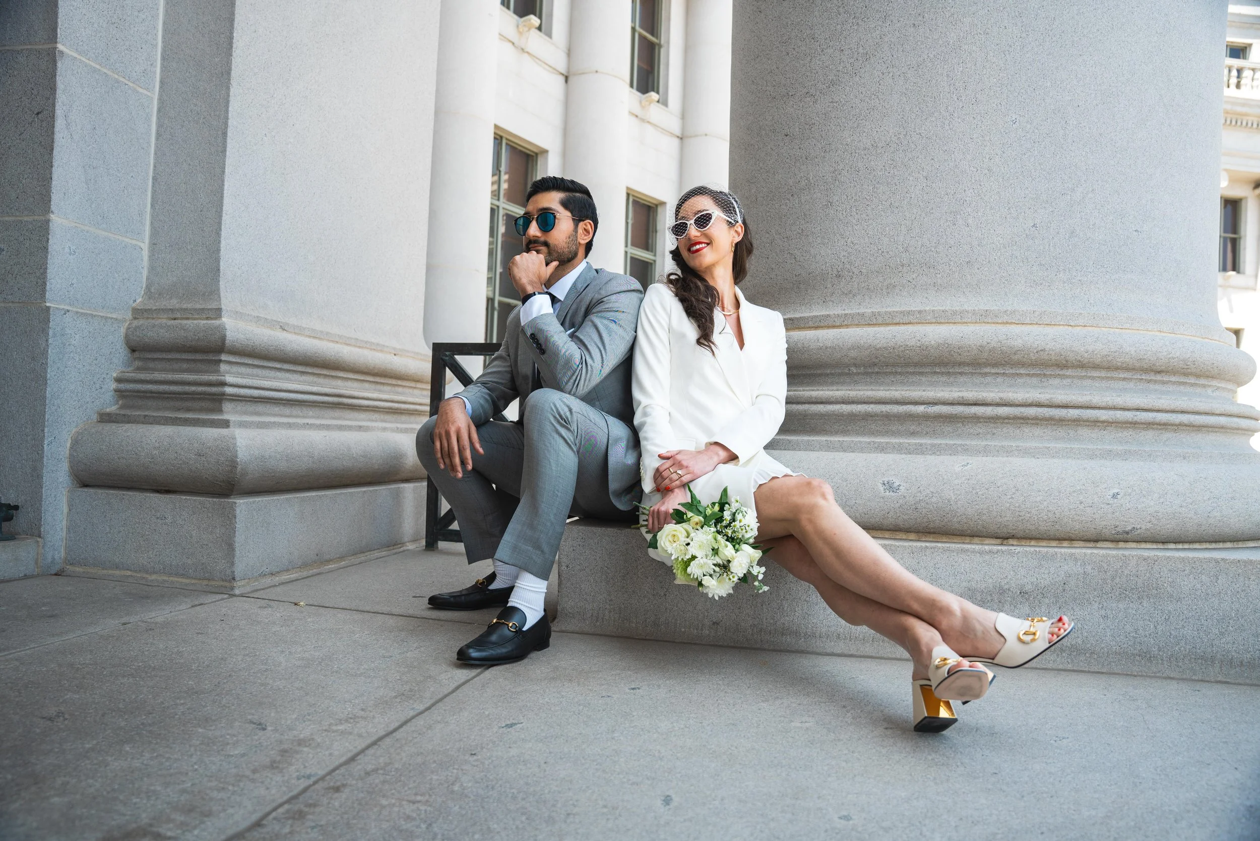 A very sharp couple in a stylized pose, waiting for the judge to arrive at the courthouse for their elopement.  The bride is wearing a bonnet, red lipstick, and heels that are flashing gold underneath.  The groom is in a sharp grey suit. 