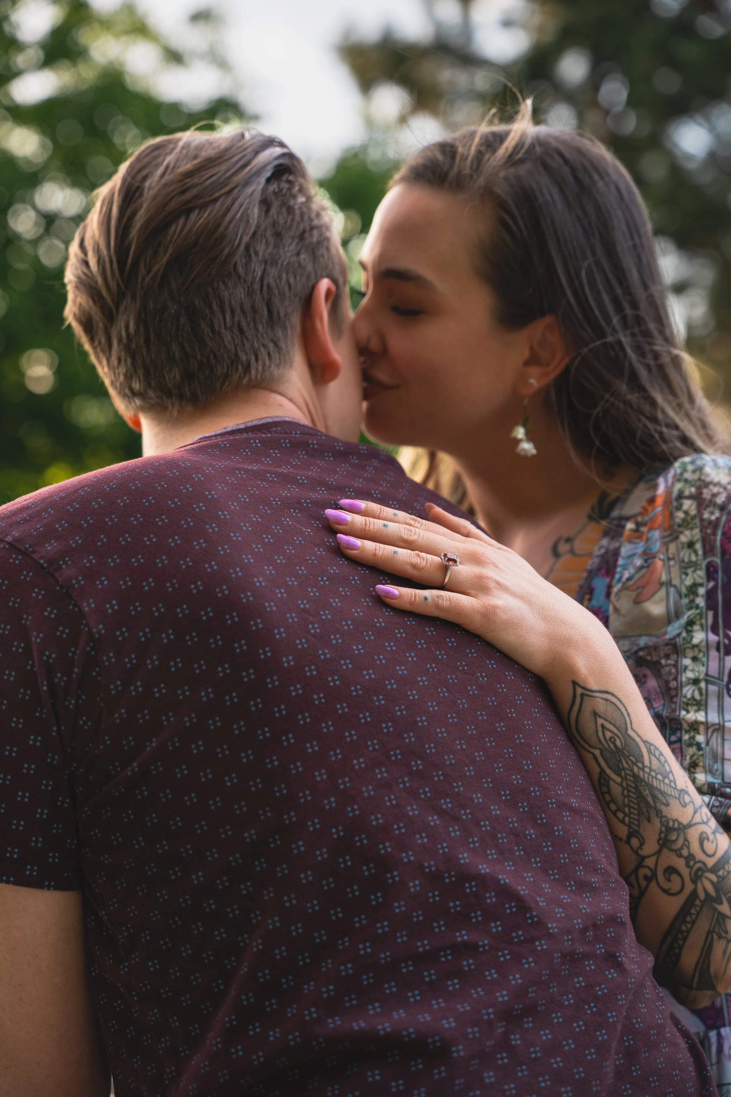 A gentle embrace between a newly engaged couple.  The girl is softly kissing the man's cheek, with her arm on his back.  Her forearm is covered in ornamental tattoos and her engagement ring is the center focus of the image.