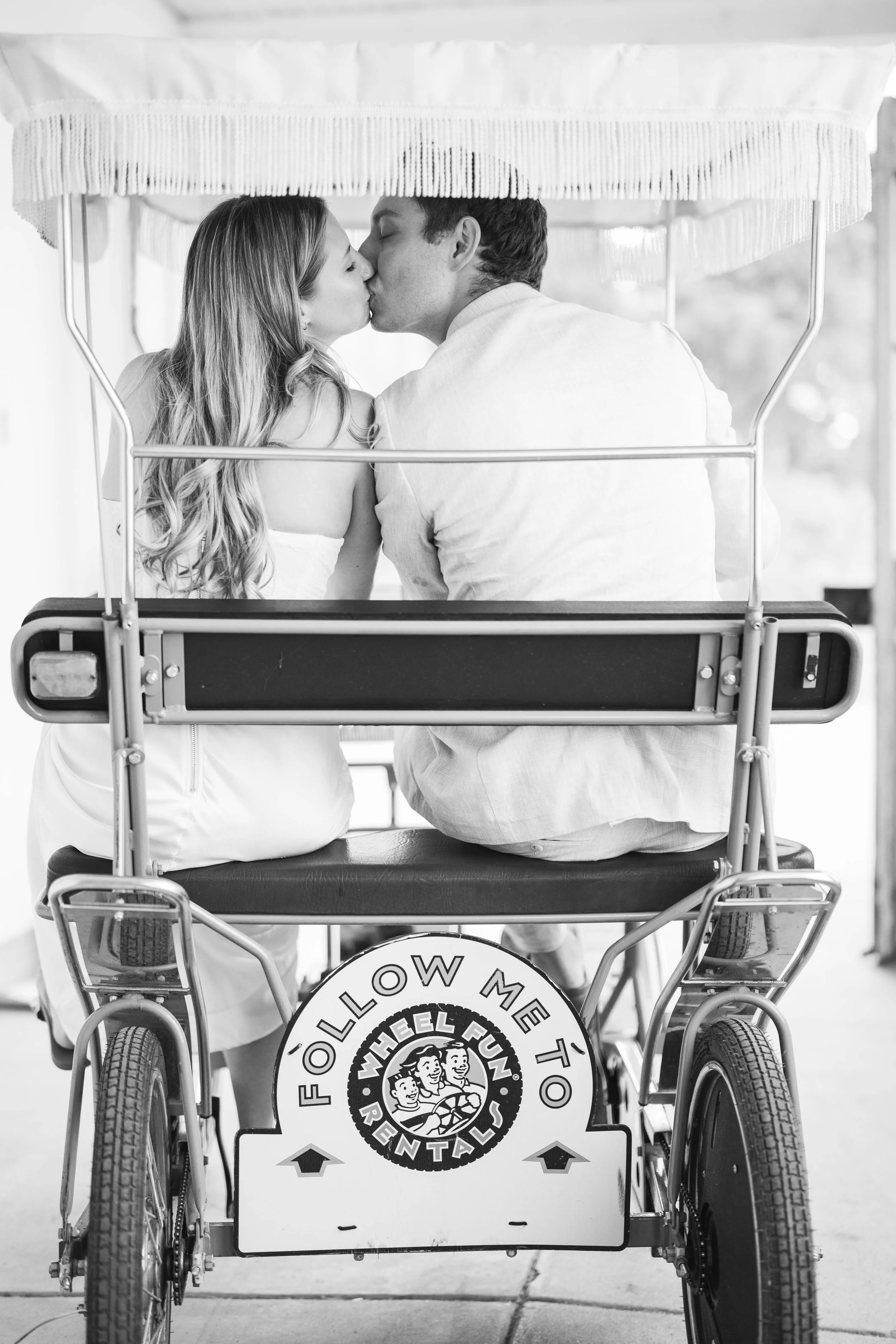 A couple kissing on a cart bicycle after eloping.  It looks like they are riding off into the sunset!