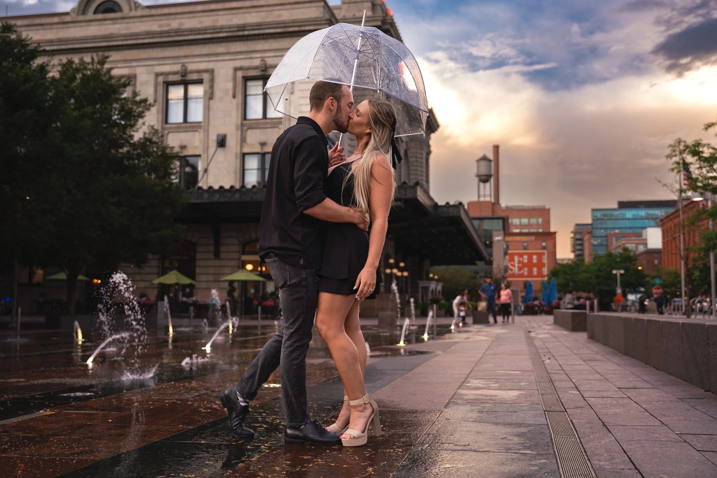 Sunset in Downtown Denver near Union Station. A water tower is in the background with golden clouds, and a couple is kissing next to a water fountain show.  He holds a clear umbrella over his head in the photo, protecting them from the rain.