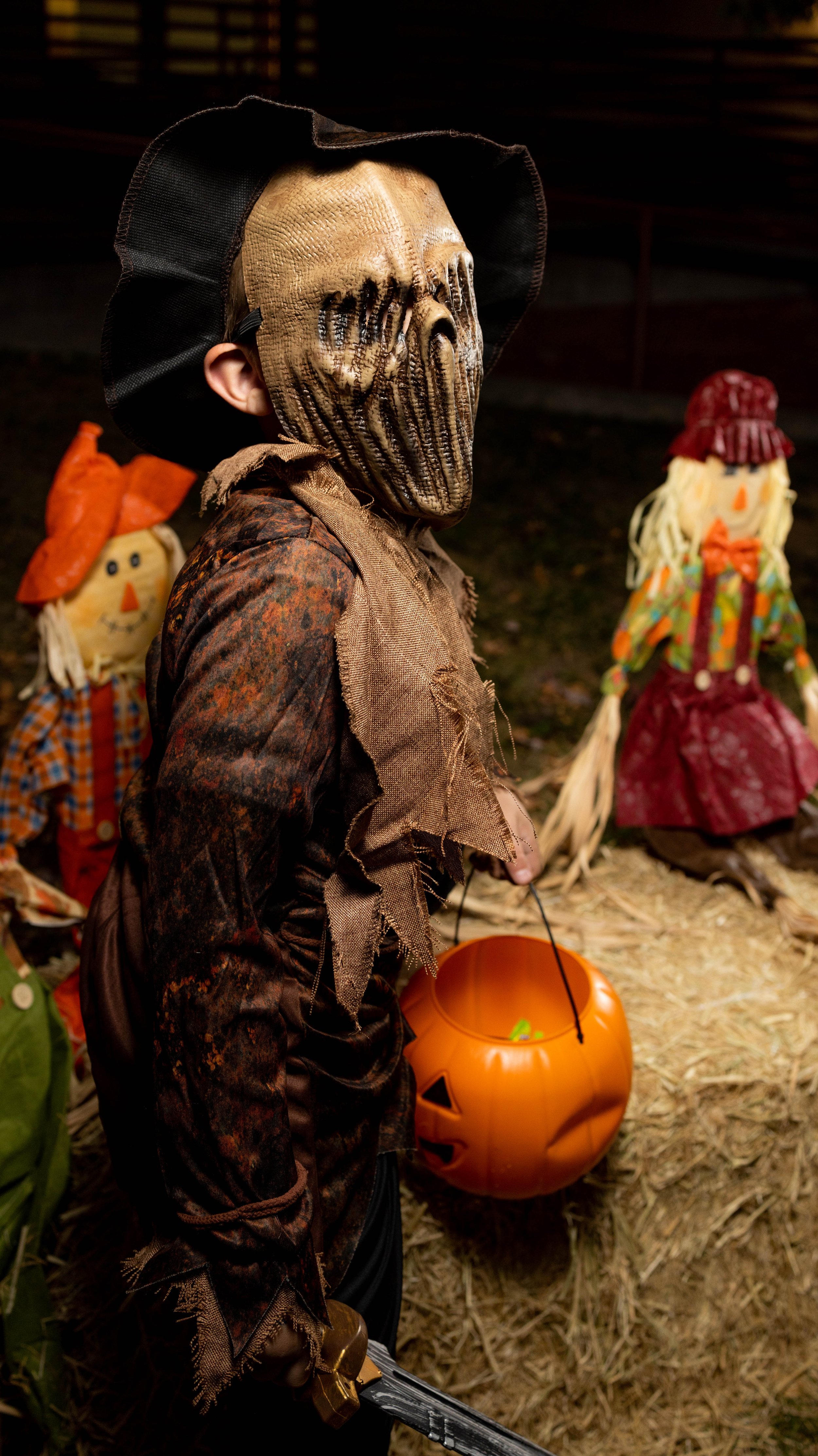 A person dressed in a Halloween costume with a scarecrow pumpkin mask, holding a plastic pumpkin bucket, standing in front of scarecrow figures at Halloween display. Photo taken by An Entropy Production.