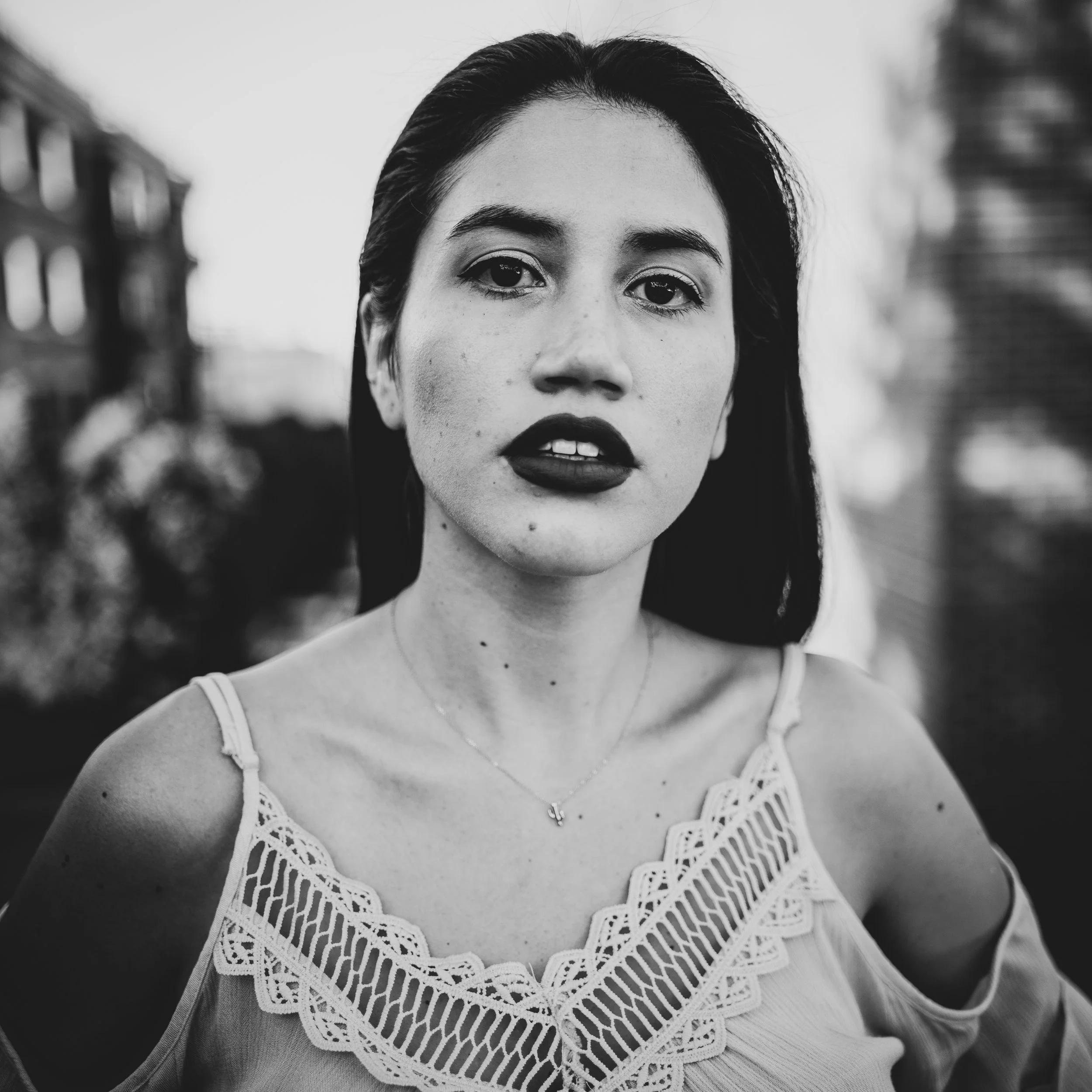 A monochrome portrait of a young woman with confident expression, wearing a lace-trimmed top in natural outdoor light. Shot by An Entropy Production, emphasizing emotion and fine detail in the subject’s face.