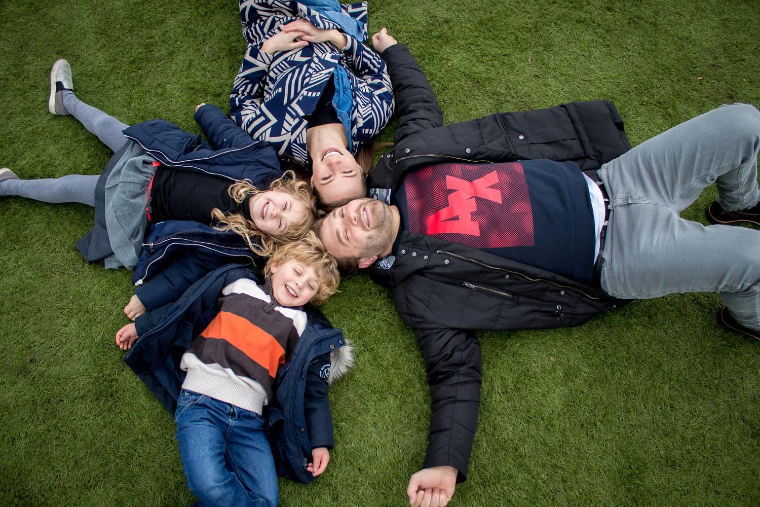 Family photography by An Entropy Production featuring a family of four lying on the grass, smiling and enjoying time together outdoors. Family photography with An Entropy Production