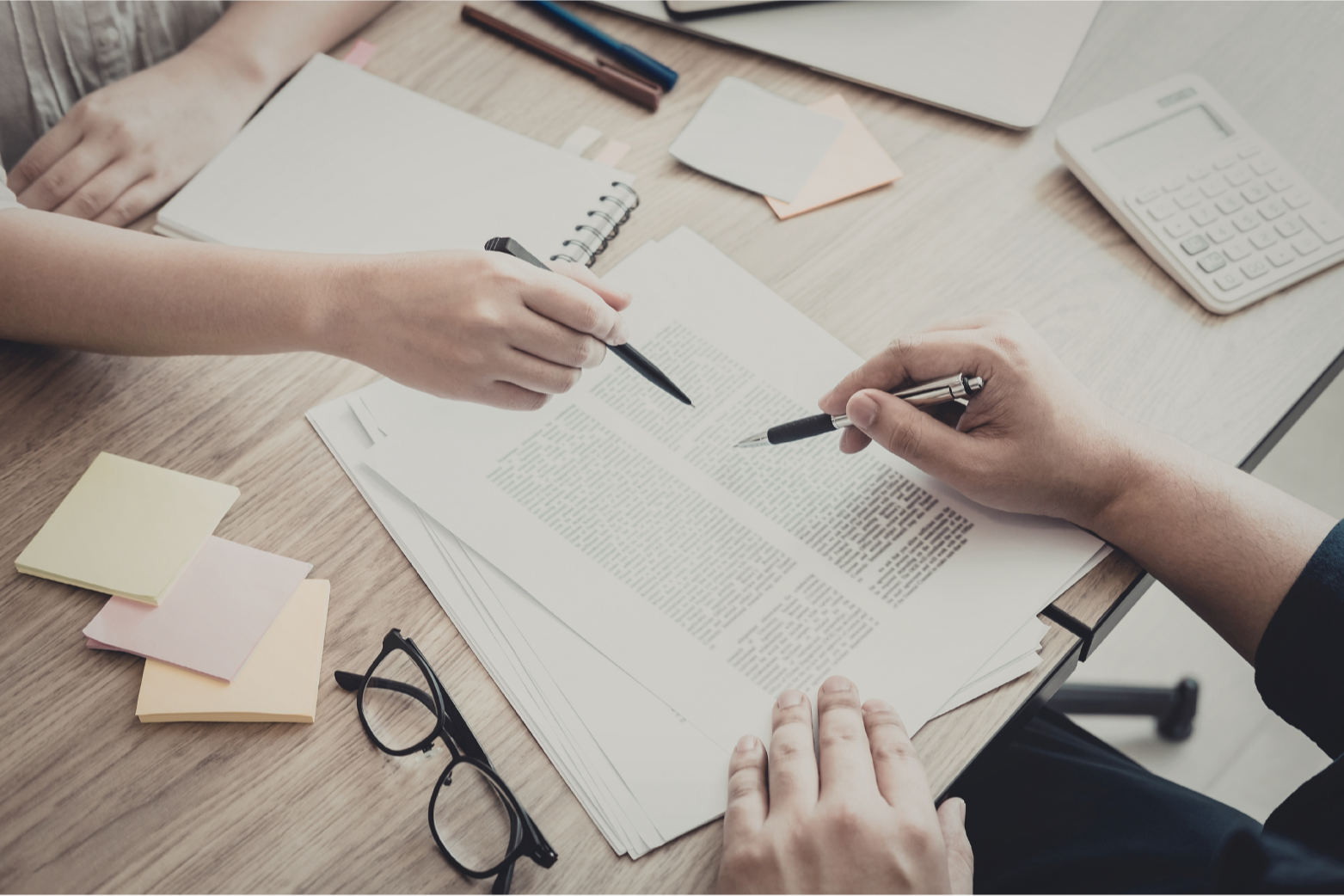Two people reviewing and discussing documents with pens, surrounded by office supplies on a wooden desk.