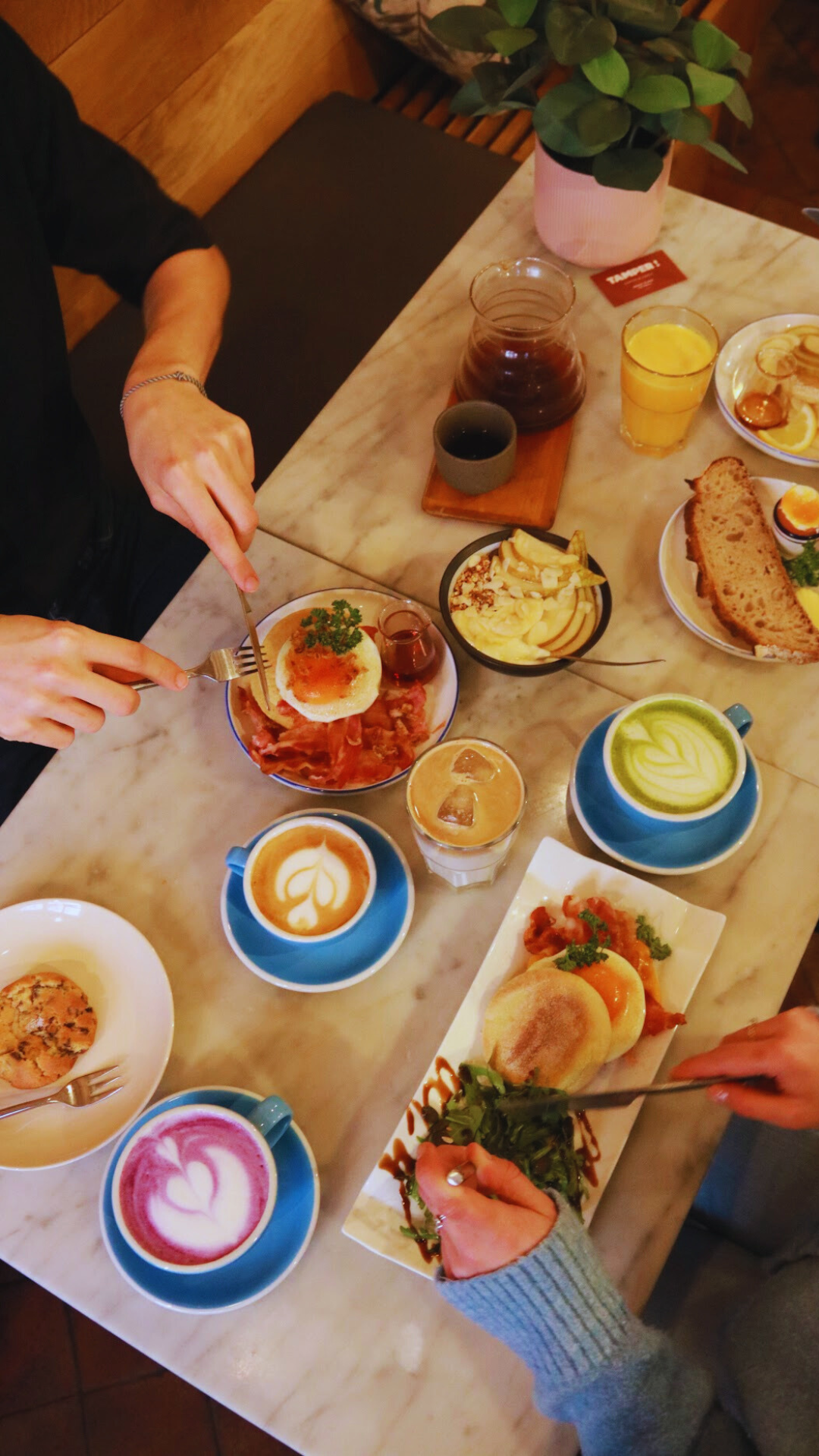 Petit déjeuner avec des cafés latte, un œuf bénédicte, un sandwich, un muffin, un gâteau, des boissons, et des plats salés sur une table en marbre.