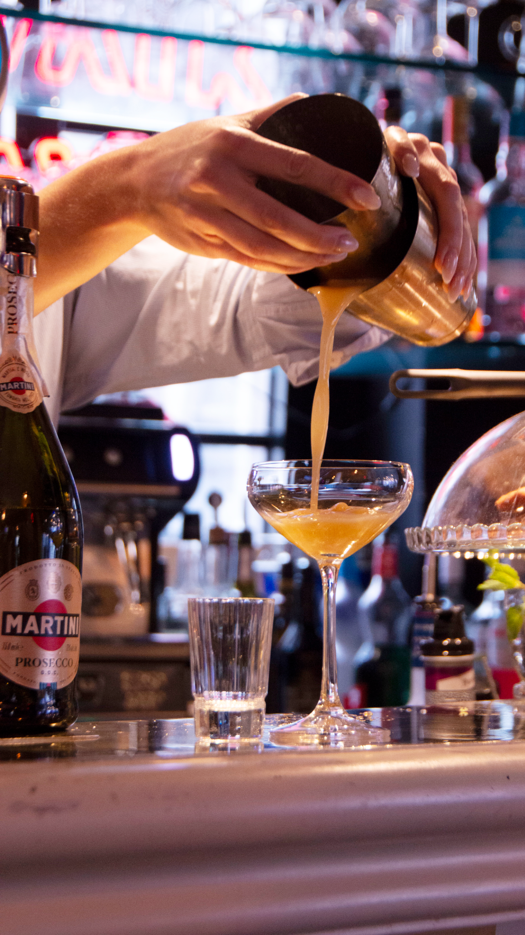 Un barman verse une boisson dans un verre à cocktail au bar. Une bouteille de Prosecco Martini est visible à gauche.