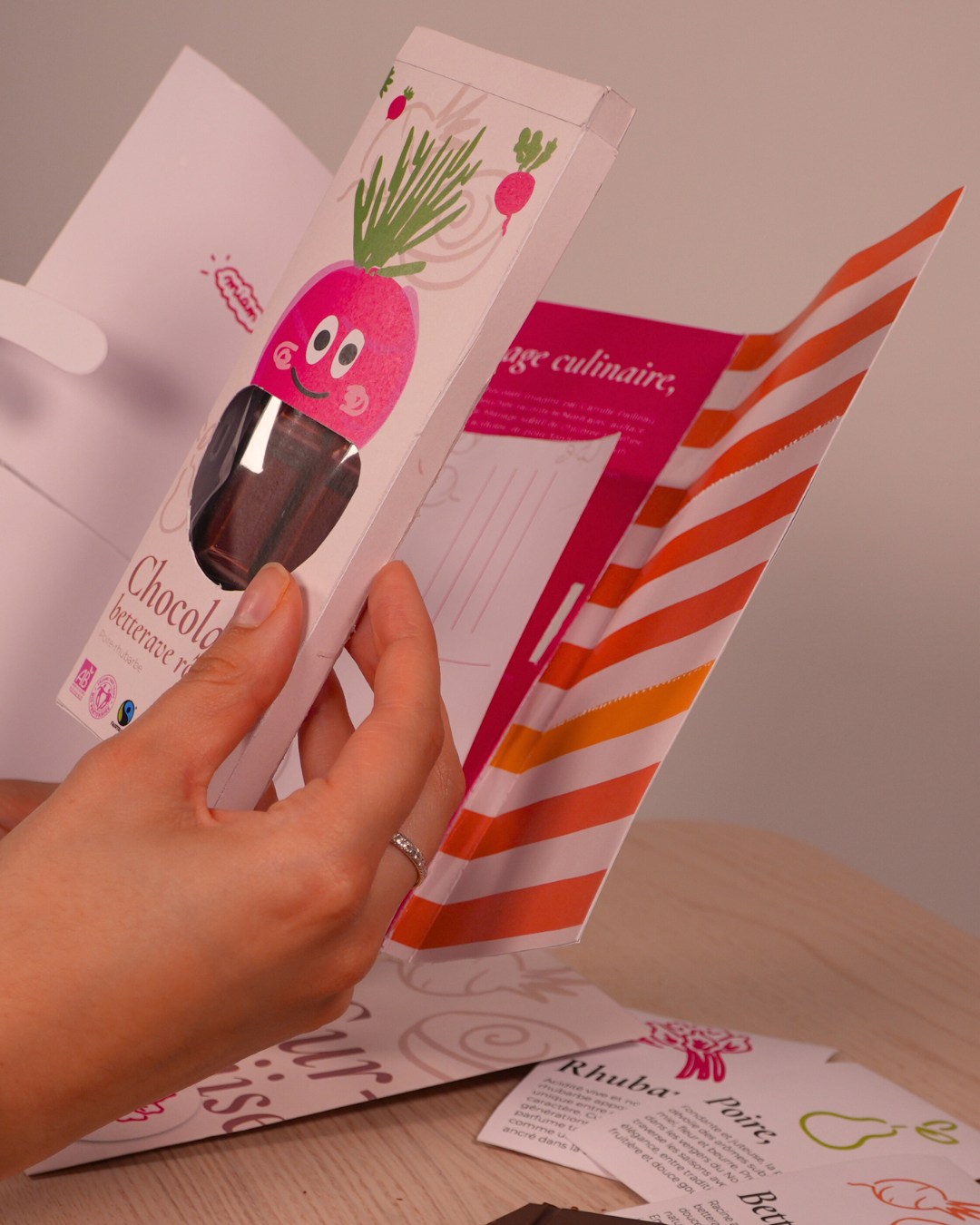 Une main tenant une boîte de chocolat avec une illustration d'une betterave souriante. La boîte est ouverte, révélant des chocolats à l'intérieur, avec d'autres documents ou cartes posés sur une table en bois.