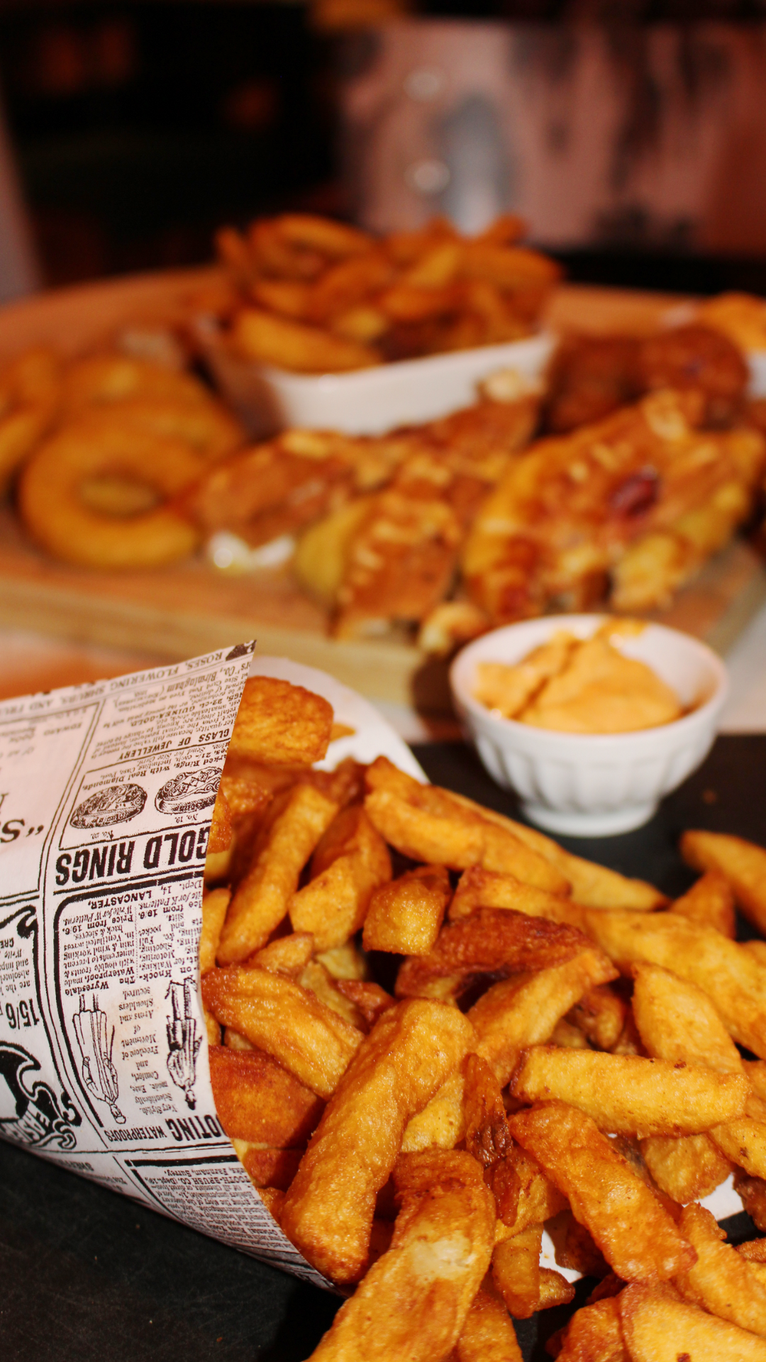 Frites dorées à l'extérieur, servies dans un papier journal imprimé, avec une sauce dans un bol blanc et des plats de poulet frit derrière.