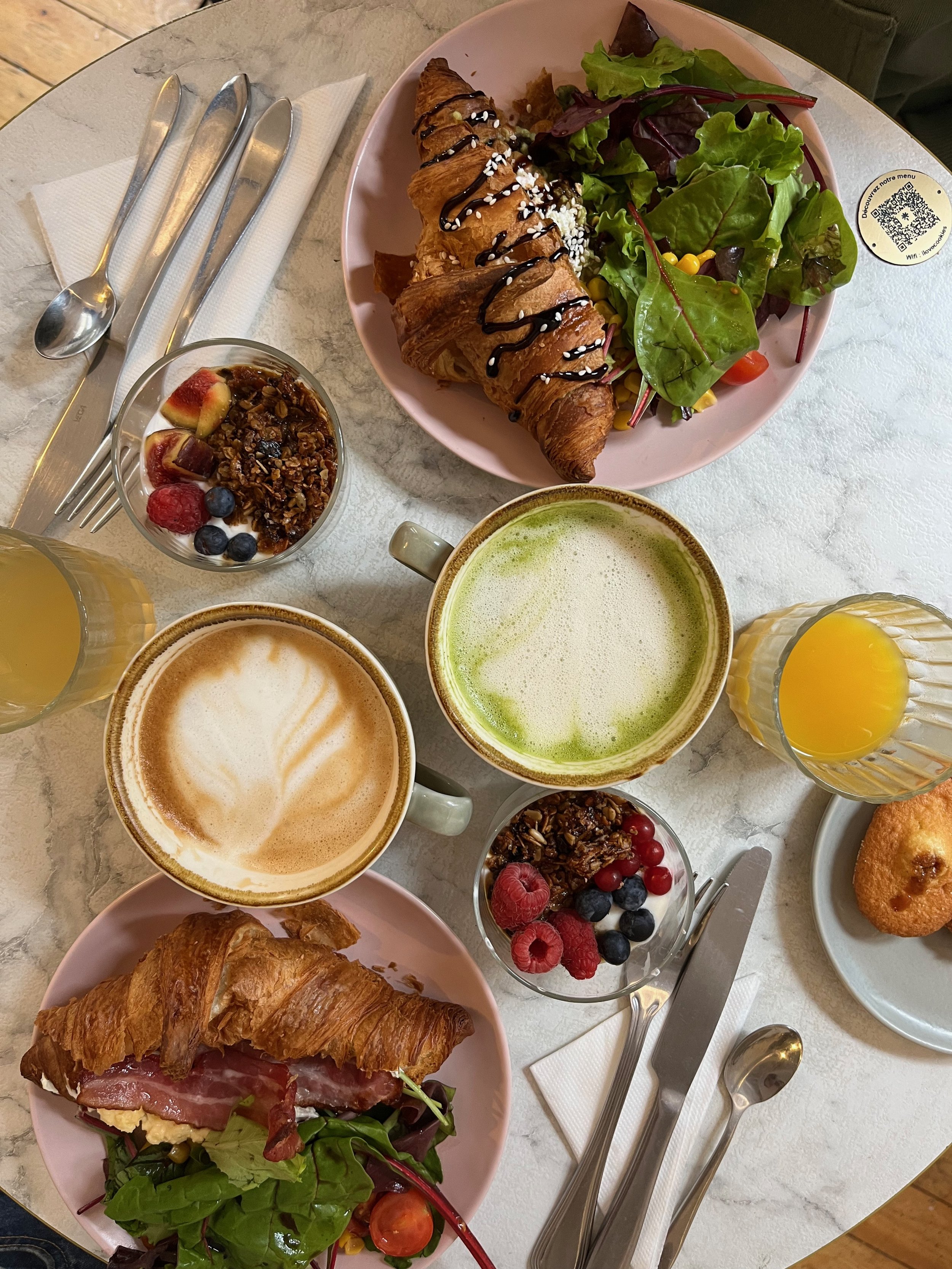 Petit-déjeuner avec croissants, salade verte, granola avec fruits, deux cappuccinos, deux jus d'orange, et une viennoiserie sur une table en marbre.