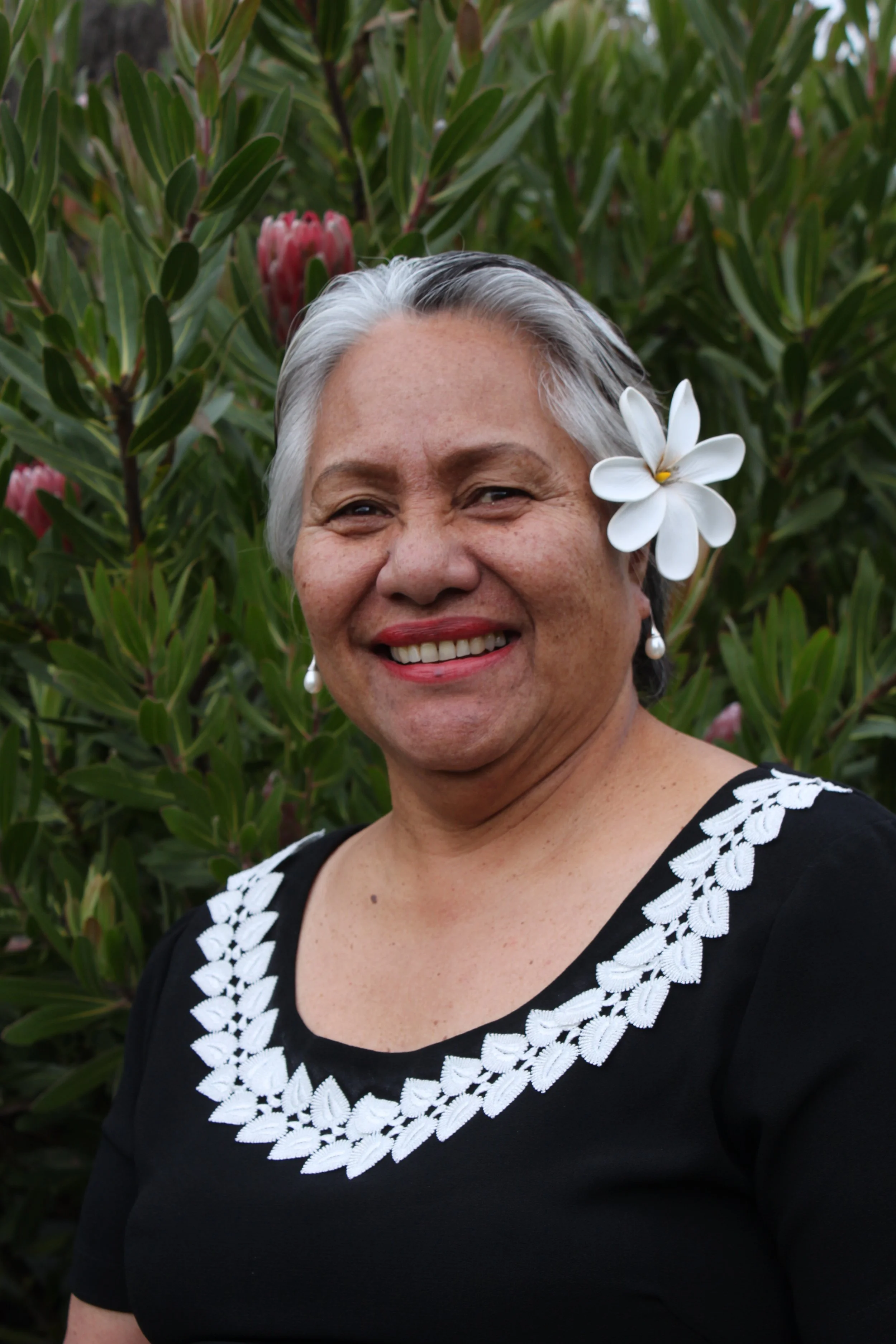 An elderly woman with gray hair, wearing a black dress with white embroidered leaf patterns along the neckline, smiles with a flower in her hair, standing outdoors among green foliage and pink flowers.