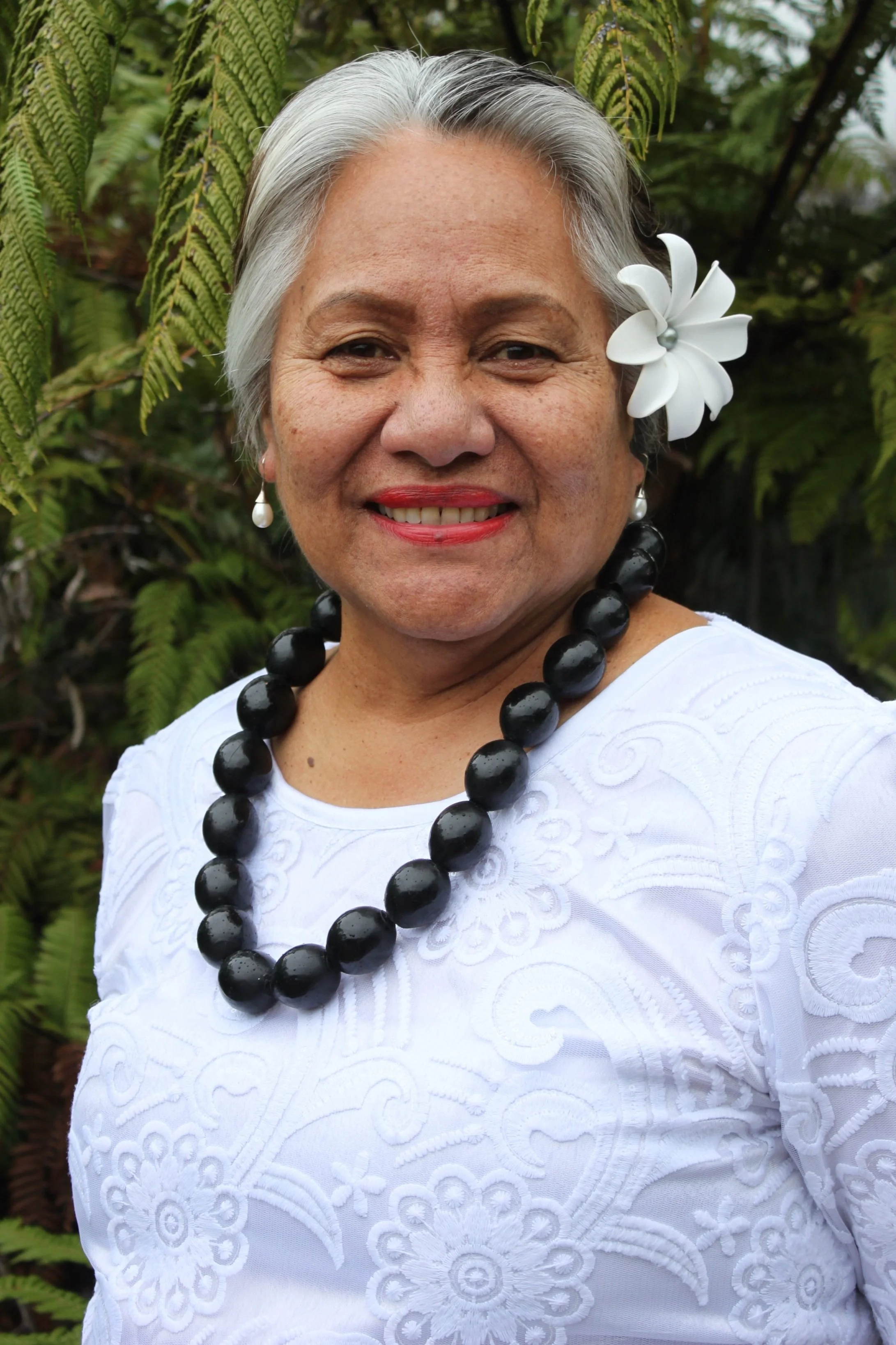 A woman with gray hair, wearing a white embroidered top, red lipstick, and pearl earrings, posing outdoors with green fern leaves in the background. She has a white flower in her hair and a large black beaded necklace.