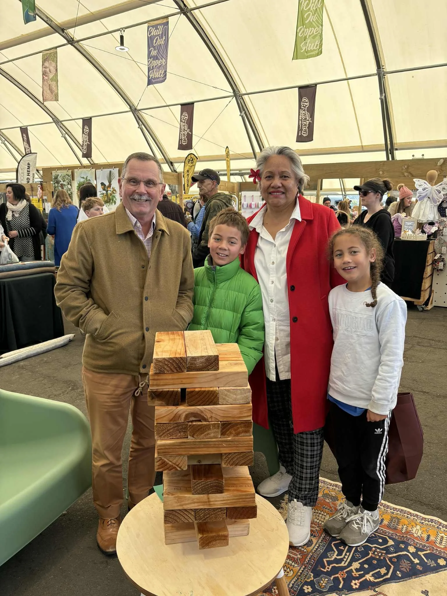 A family of four standing around a large wooden Jenga game inside a spacious tent at an indoor marketplace or fair. The family includes a man, woman, and two children, with several other shoppers and booths visible in the background.