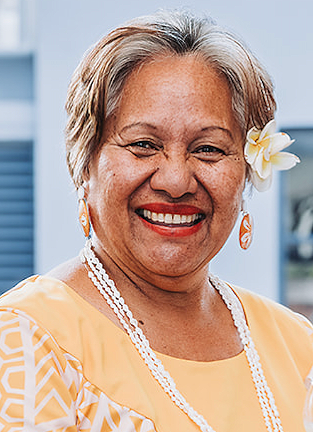 Smiling older woman with short gray hair, wearing a yellow dress with white patterns, a flower in her hair, and pearl jewelry, outdoors with a blue building in the background.
