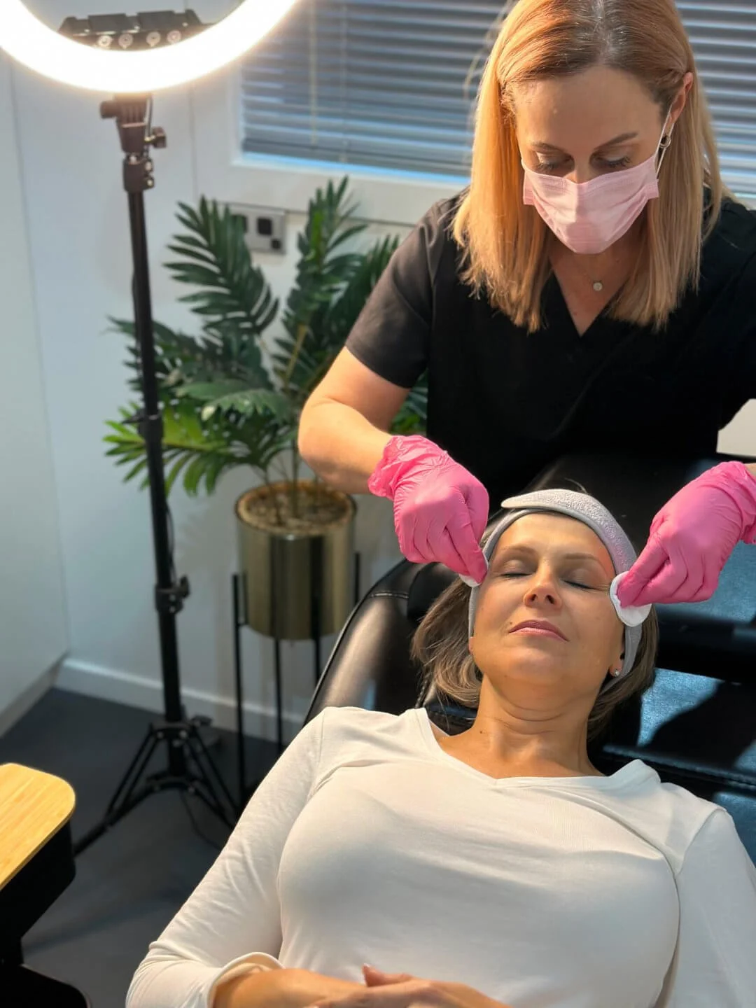 A client lying on a treatment bed with a towel around her head at the clinic, receiving a facial treatment from RN Jolieta wearing pink gloves and a pink mask, with a large ring light and a green plant in the background.