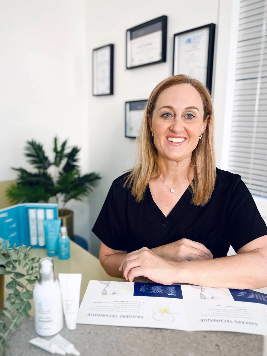 RN Jolieta is wearing a black scrub top, sitting at a desk with skincare products and informational pamphlets, in the clinic with framed certificates on the wall and a small potted plant in the background.
