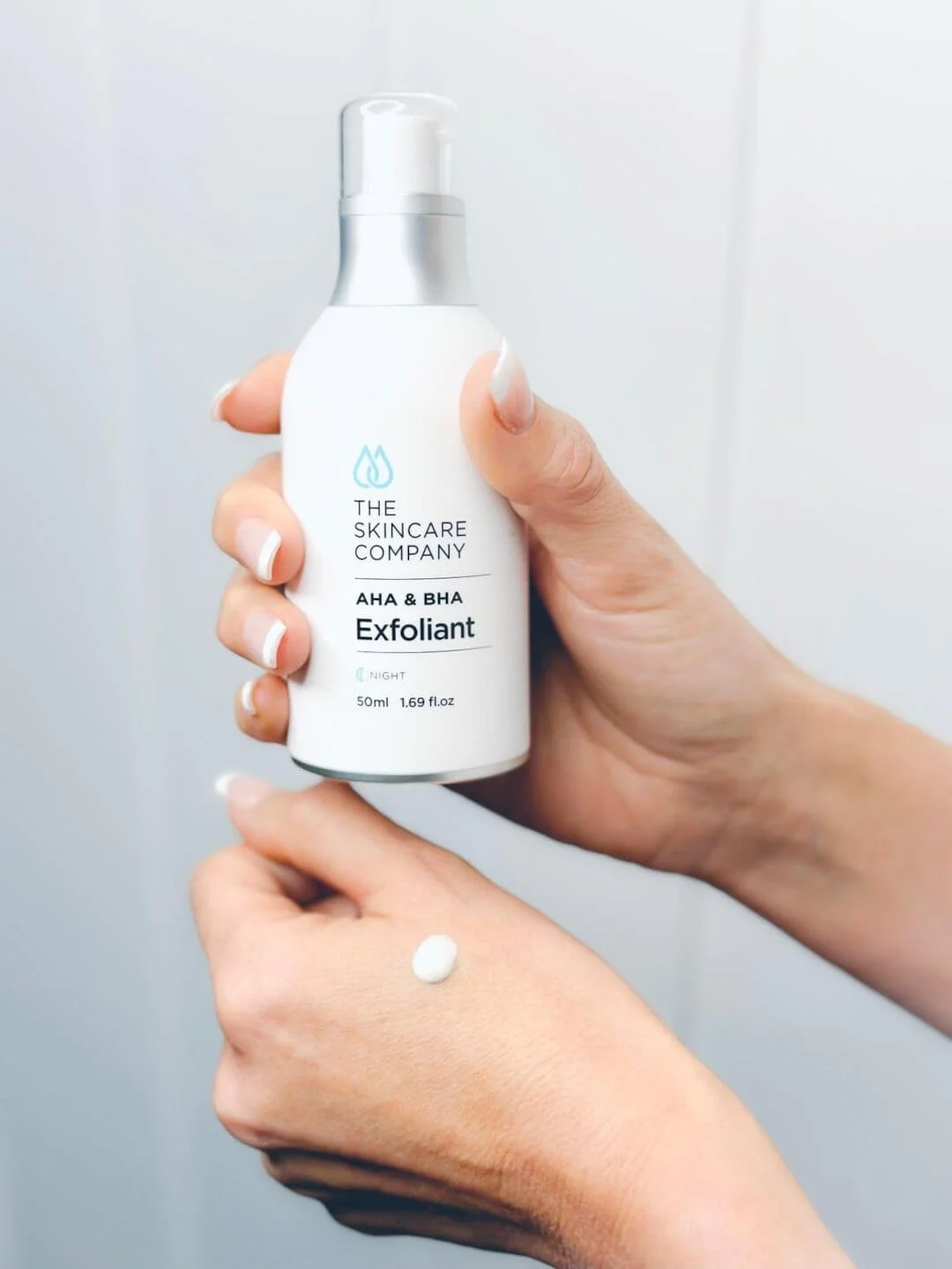 Hand holding a white bottle of skincare exfoliant labeled 'The Skincare Company AHA & BHA Exfoliant Night' with a small dollop of the product on the hand.